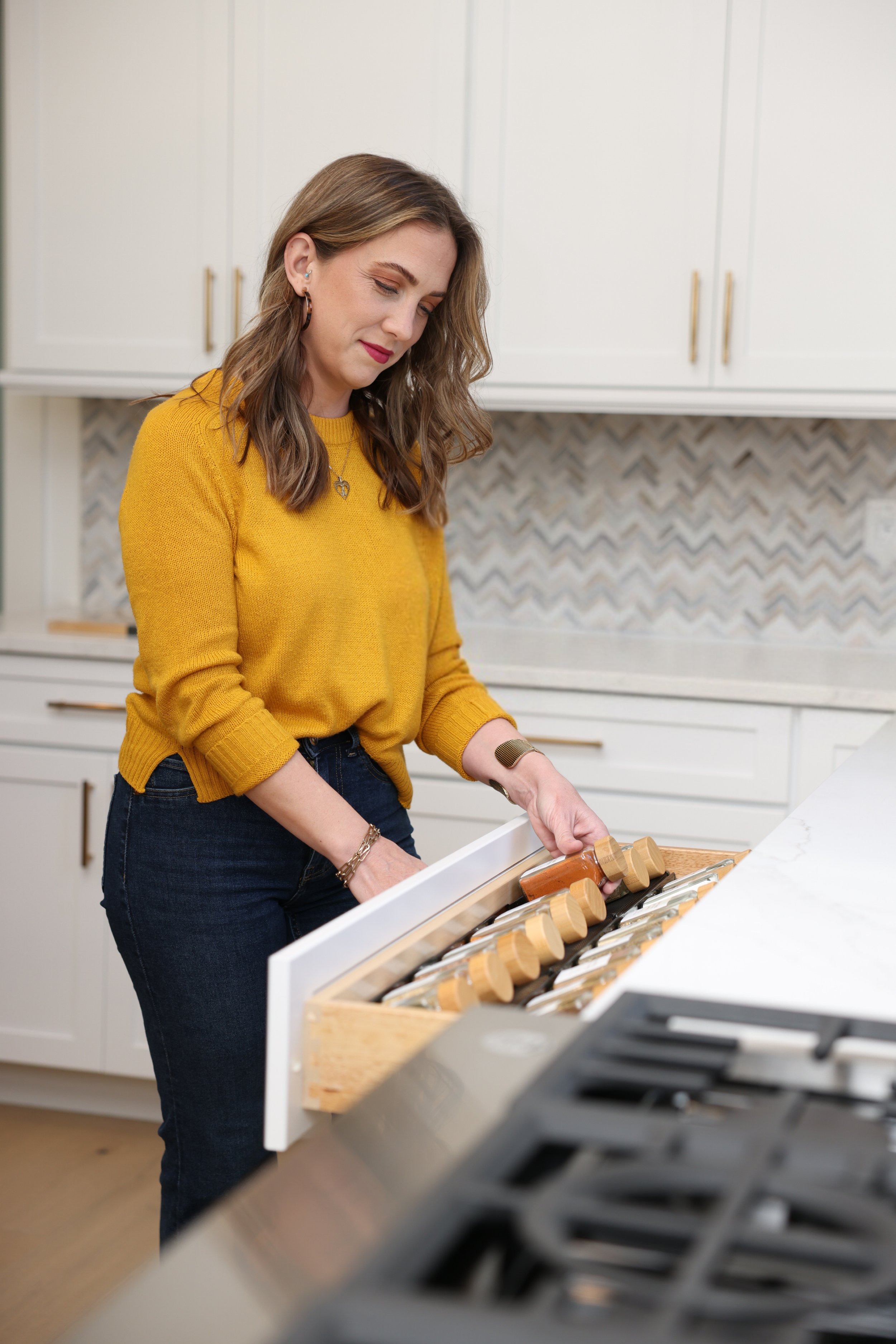 A woman wearing a yellow sweater and dark jeans is in a modern kitchen, opening a wooden drawer with wine bottles inside, near a white countertop and black kitchen appliances.