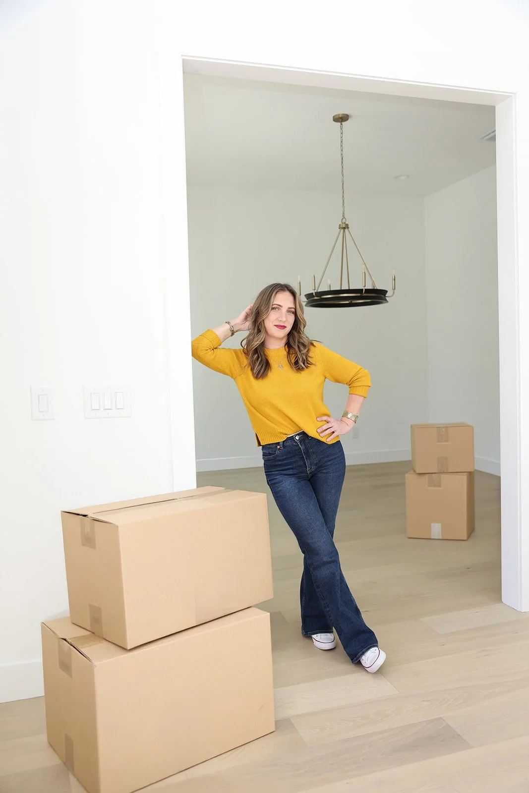 A woman in a yellow sweater and jeans standing in an empty house with moving boxes.