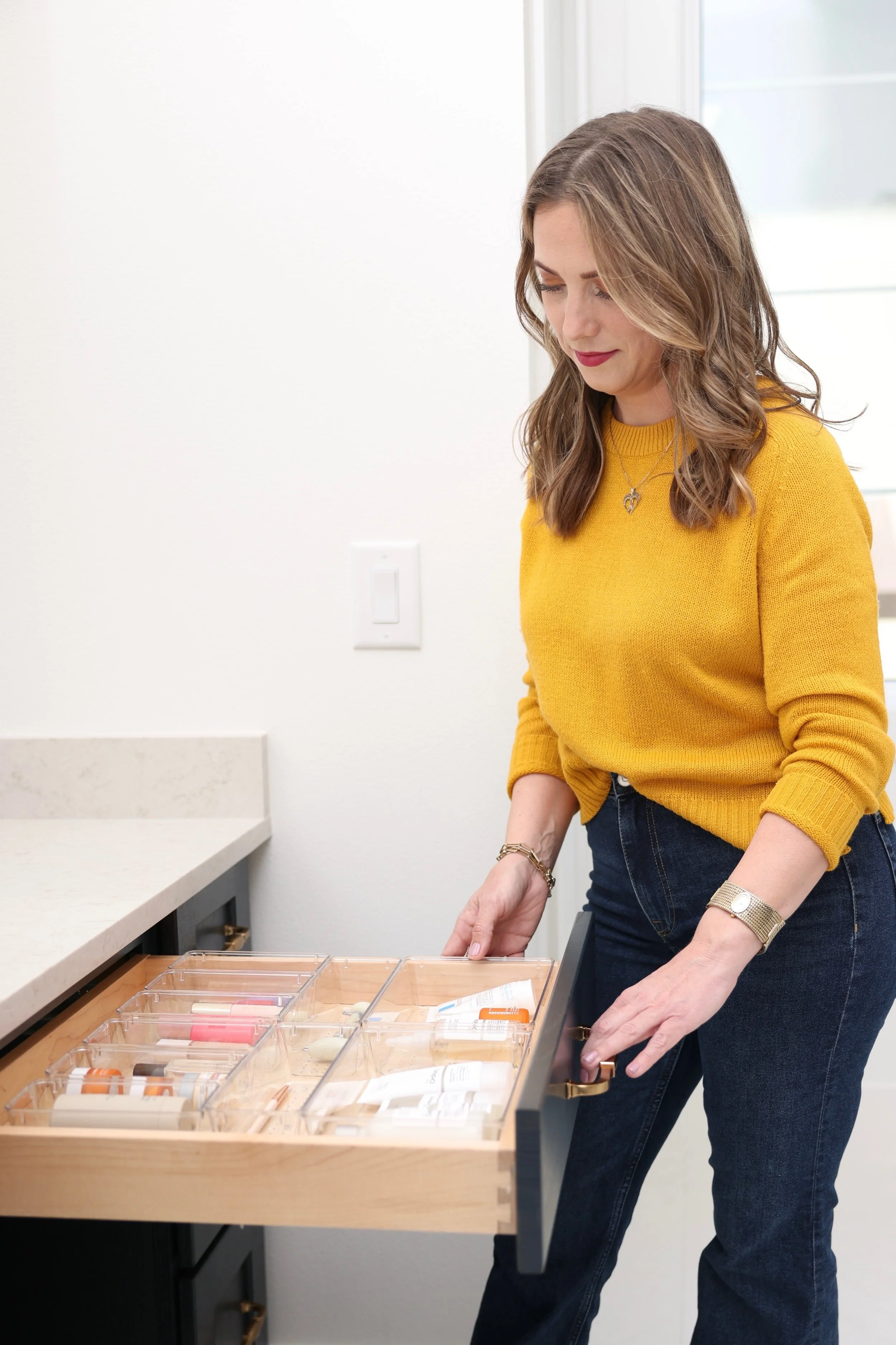 Woman in yellow sweater organizing cosmetics or toiletries in a drawer at a bathroom vanity.