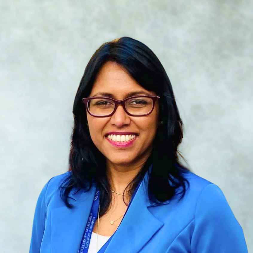 A woman with shoulder-length black hair, wearing glasses, a blue blazer, and a white top, smiling in front of a plain background.
