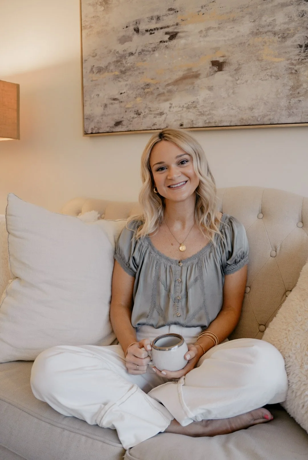 Ashton landers sitting cross-legged on a beige sofa, holding a mug, with a large abstract painting behind her and pillows on her sides.