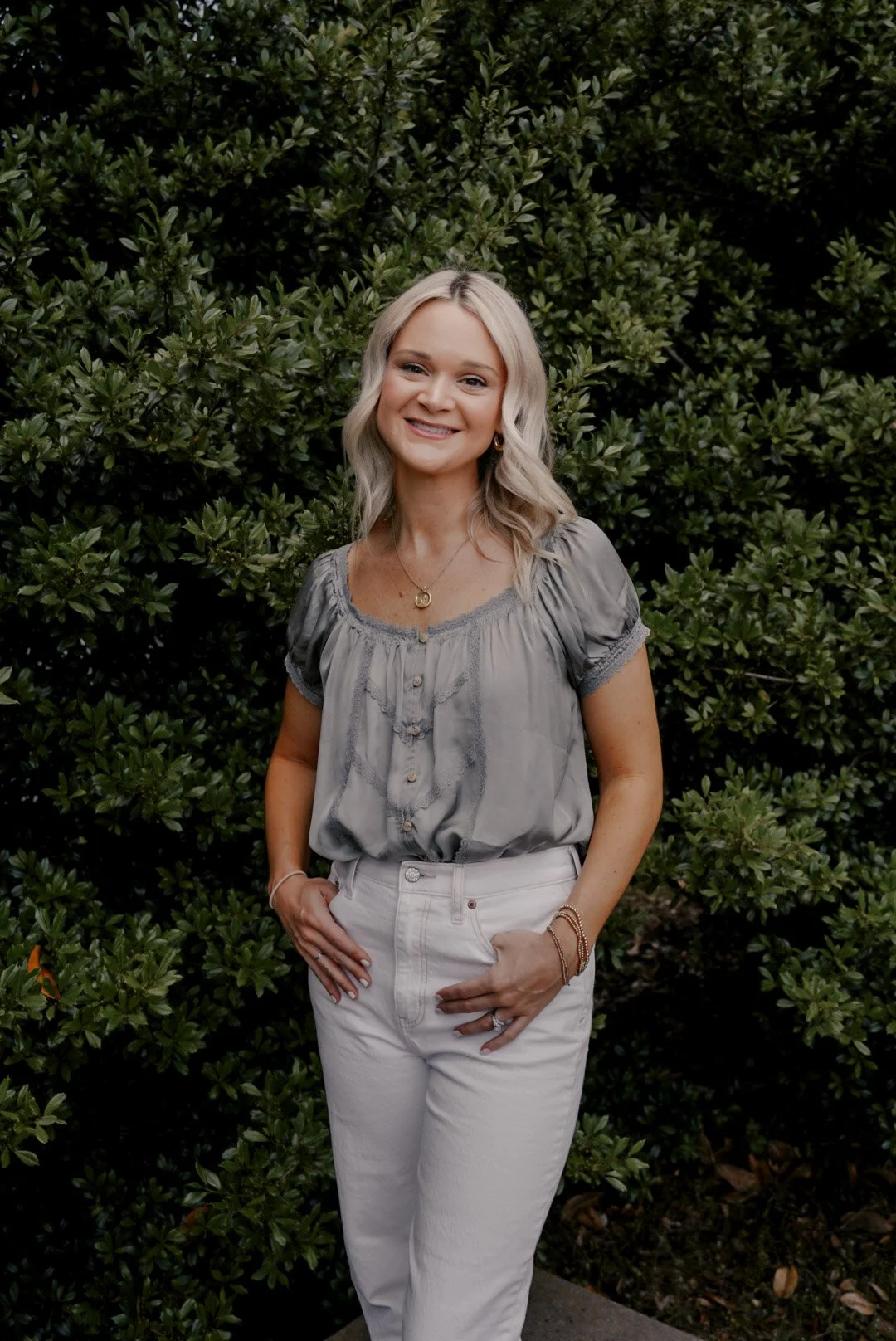 Ashton's headshot, wearing a gray blouse and white pants, standing in front of green bushes.
