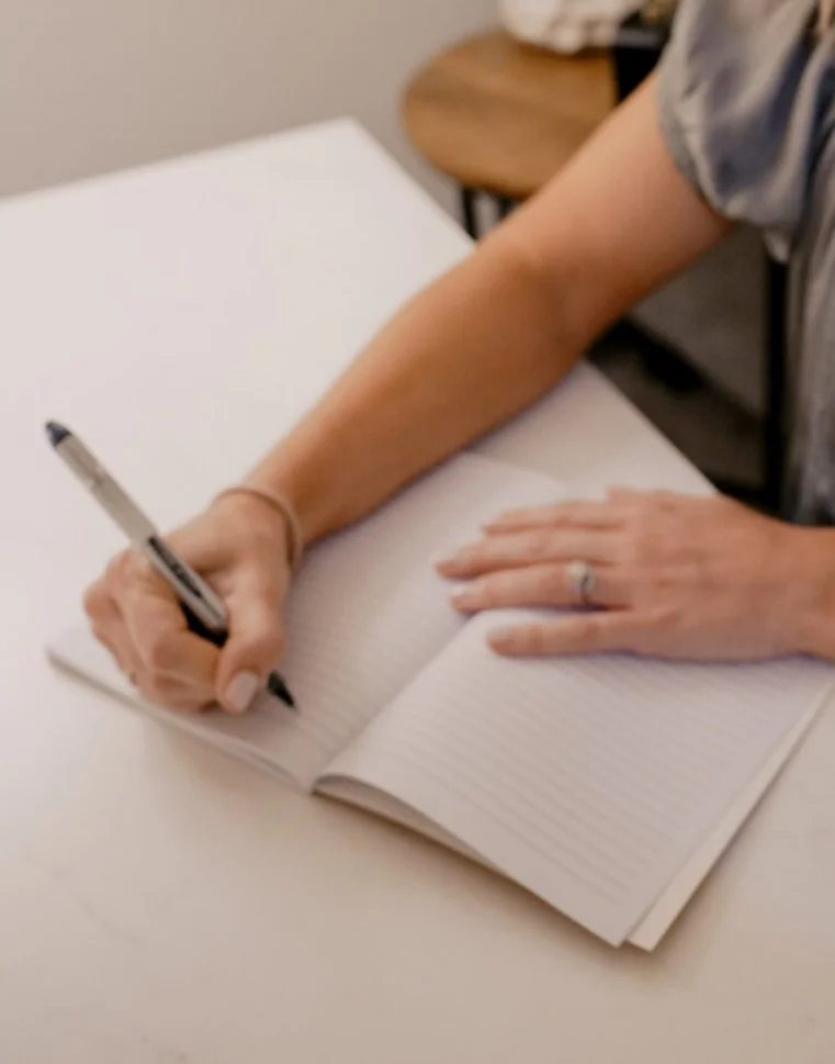 Close up shot of Ashton writing in a lined notebook with a black pen, sitting at a white table.