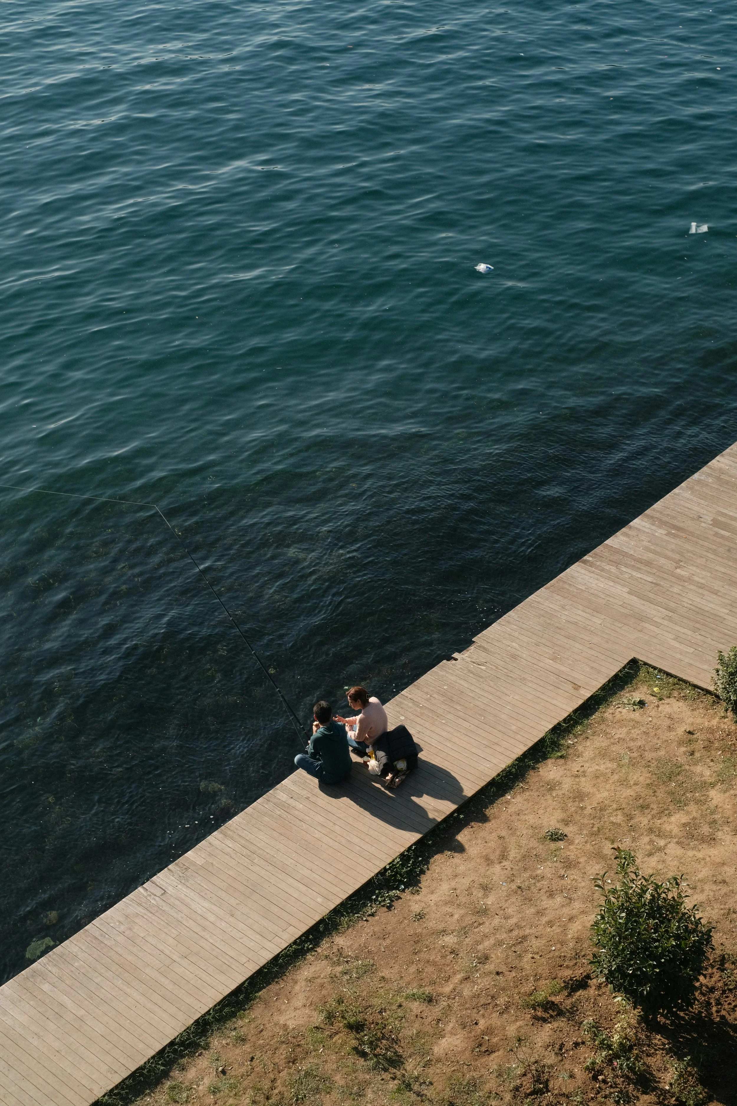 Two people sitting on a wooden dock by a body of water, fishing. Part of the dock and some grass and small bushes are visible in the foreground.