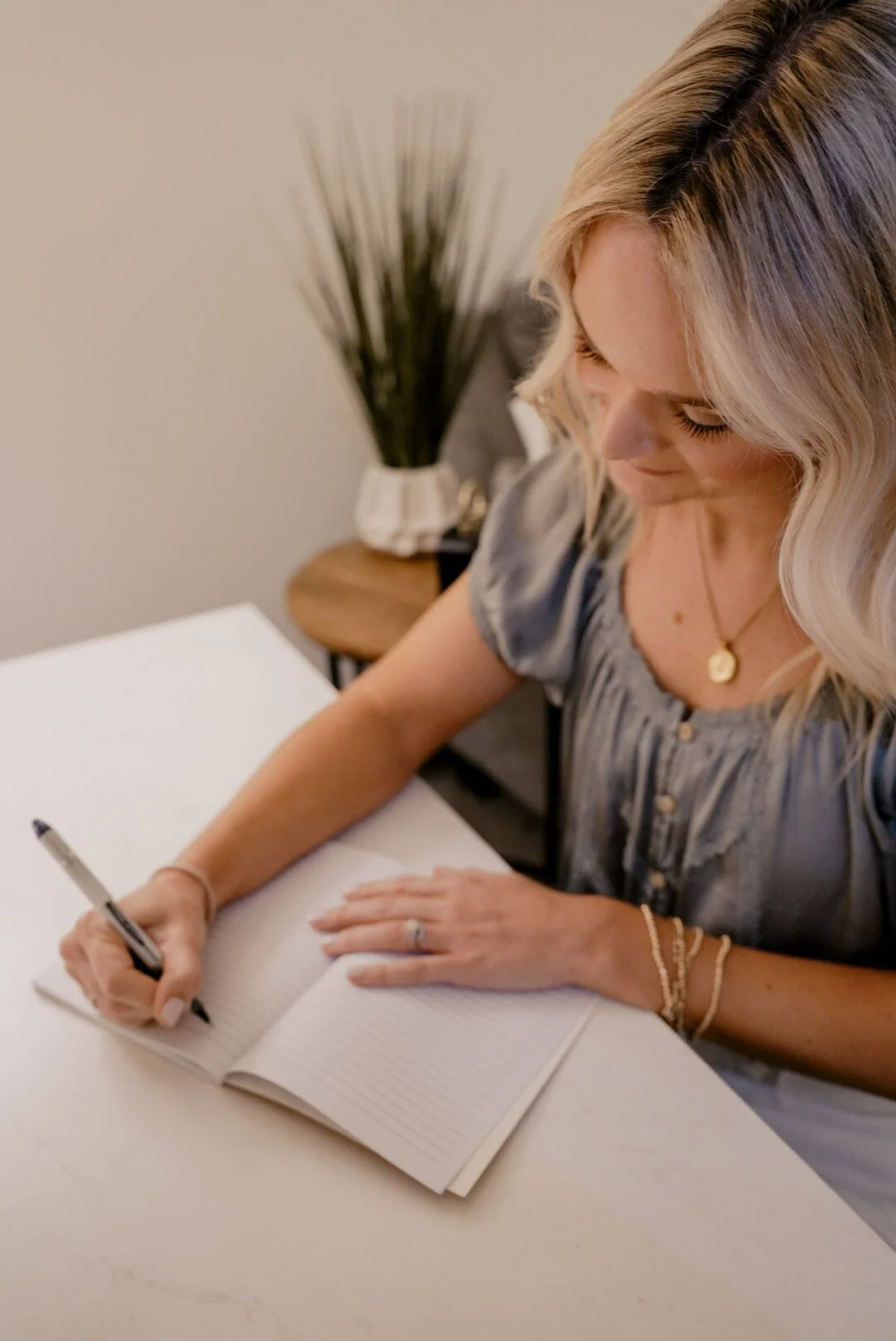 Ashton writing in a lined notebook at a white table, with a side table holding a decorative plant in the background.