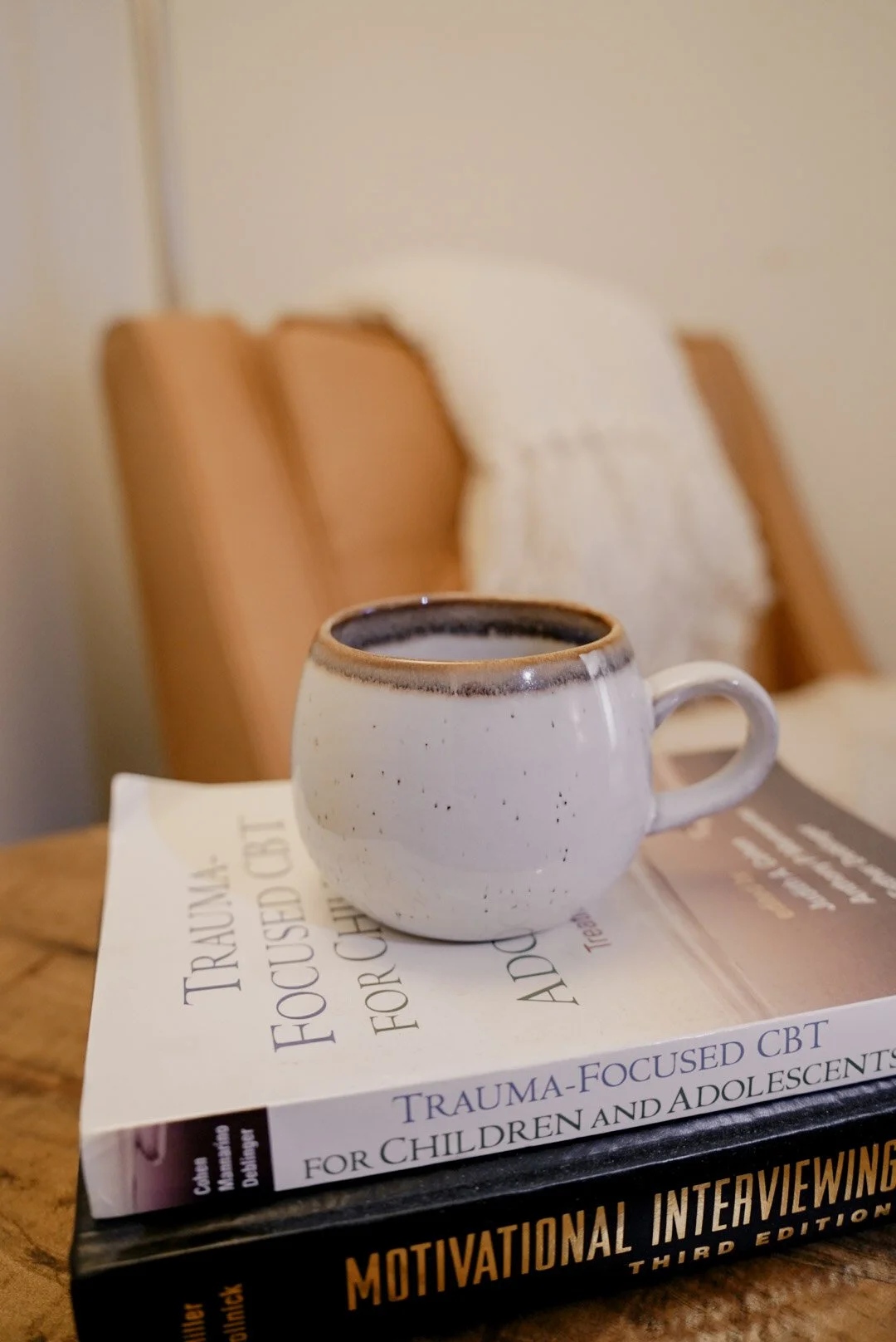 A white ceramic mug with a dark rim on a book titled "Trauma-Focused CBT for Children and Adolescents." The mug is on a wooden surface, with a beige cushion and chair in the background.