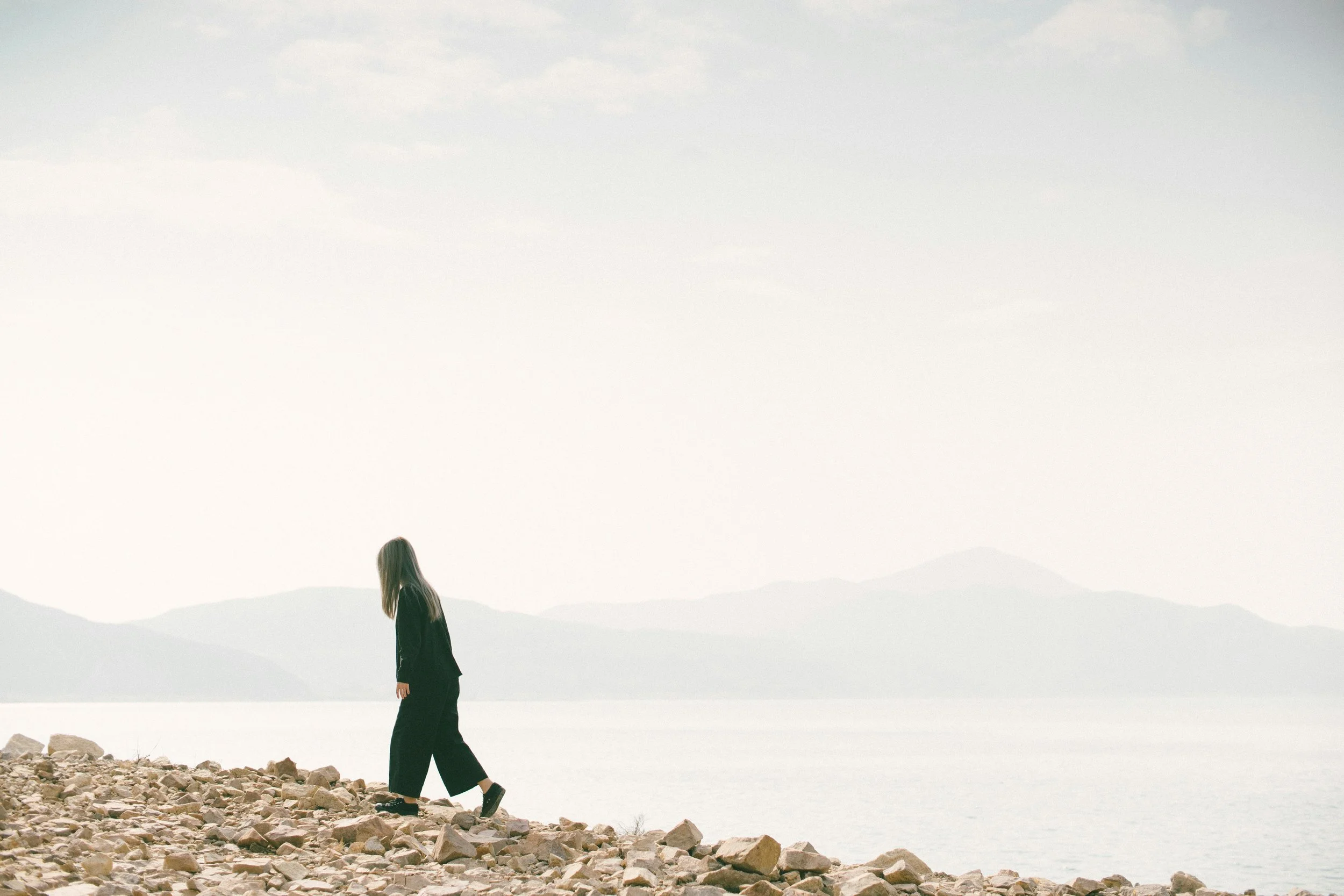 A woman with long blonde hair, dressed in black, walking along a rocky shoreline near a body of water with mountains and a cloudy sky in the background.