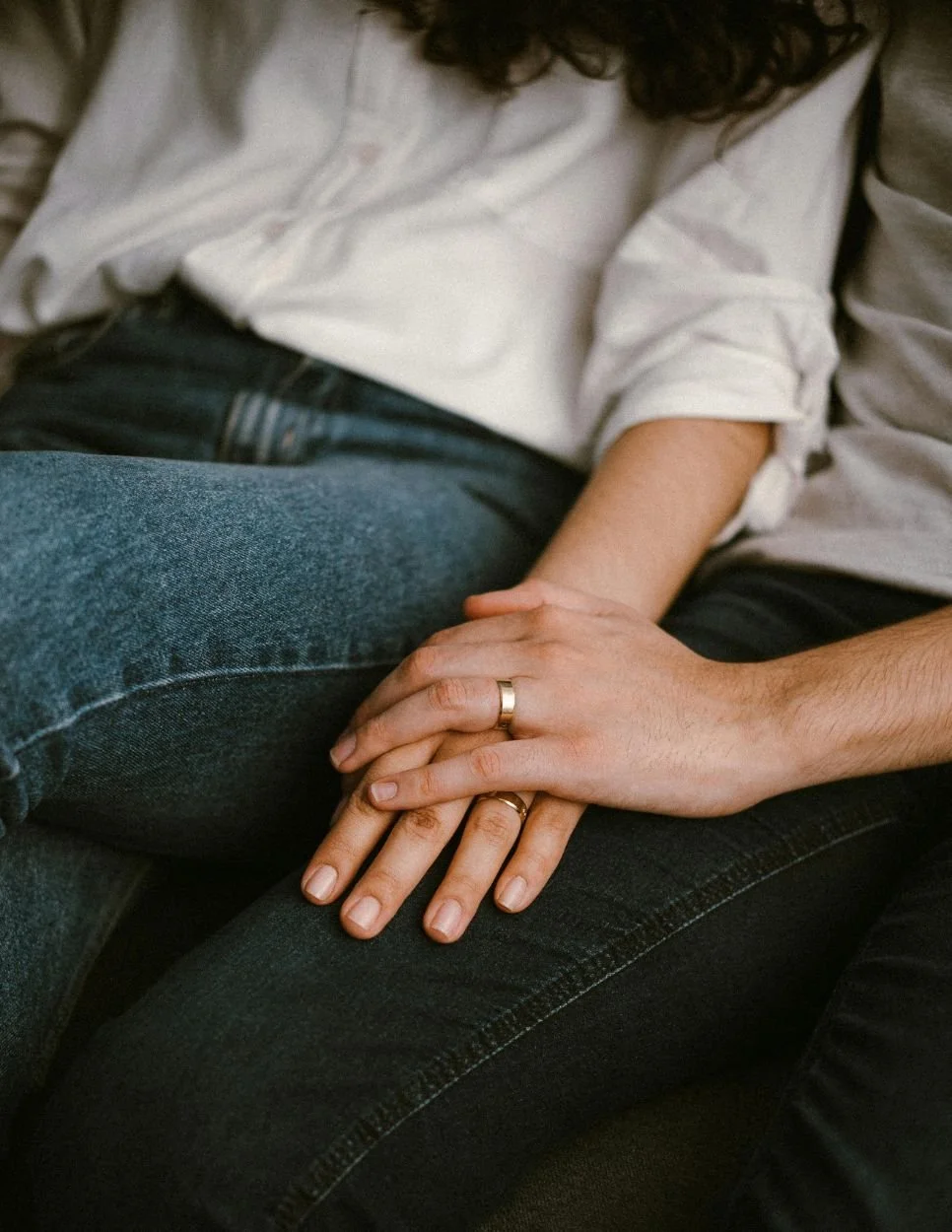 A couple holding hands, with wedding bands, sitting close together on a sofa.