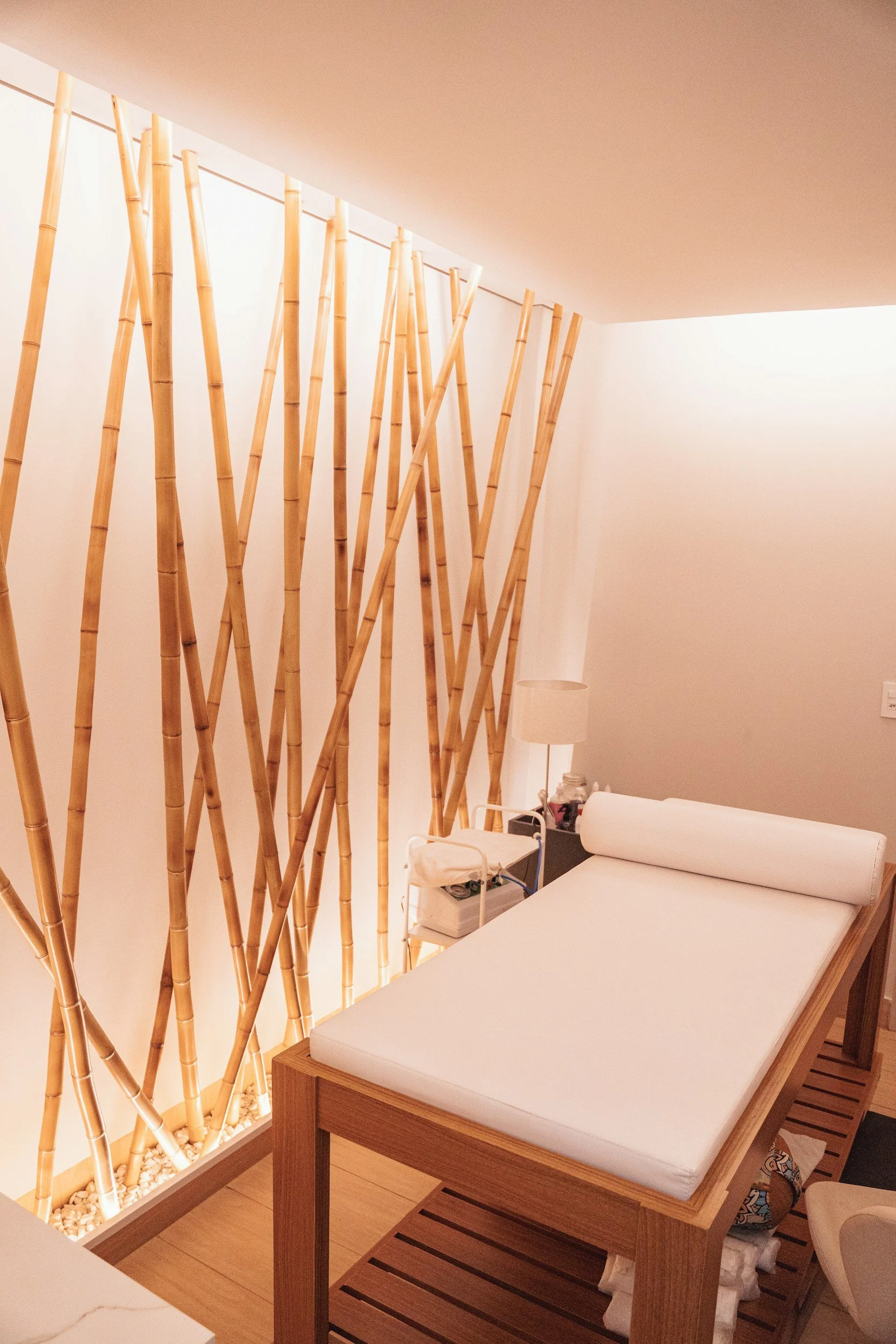 A spa or massage room with a bamboo decorative wall partition, a massage table with white linens, a bedside lamp, and a small table with various items.