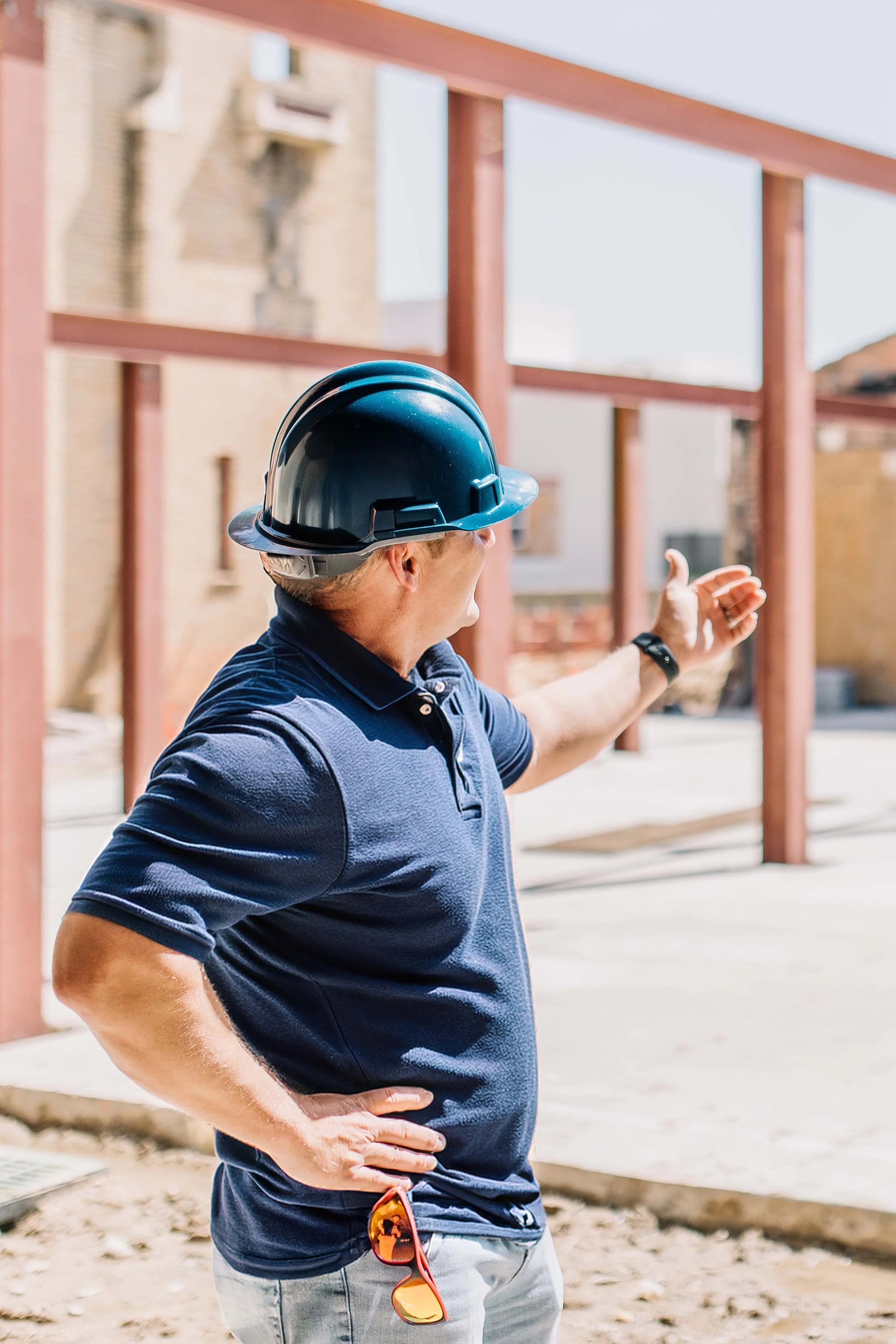 A man wearing a black construction helmet, a blue polo shirt, and sunglasses hanging from his waistband, standing outdoors at a construction site with his hand on his hip and the other arm extended, pointing at something.