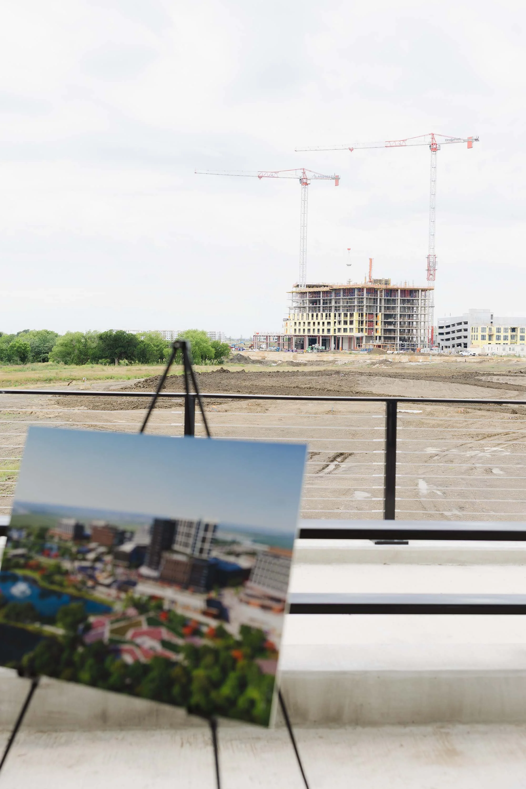 Construction site with two cranes and a building under construction, view from indoors with blurred image of buildings and trees in the foreground.