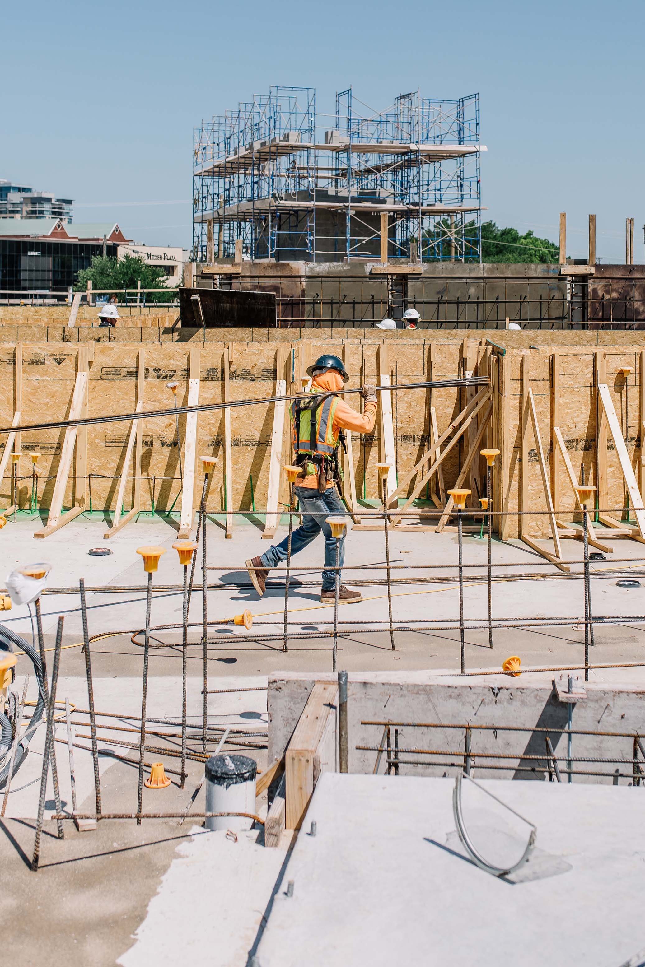 Construction site with workers wearing safety helmets and orange safety vests, wooden frameworks, concrete foundation, and scaffolding.