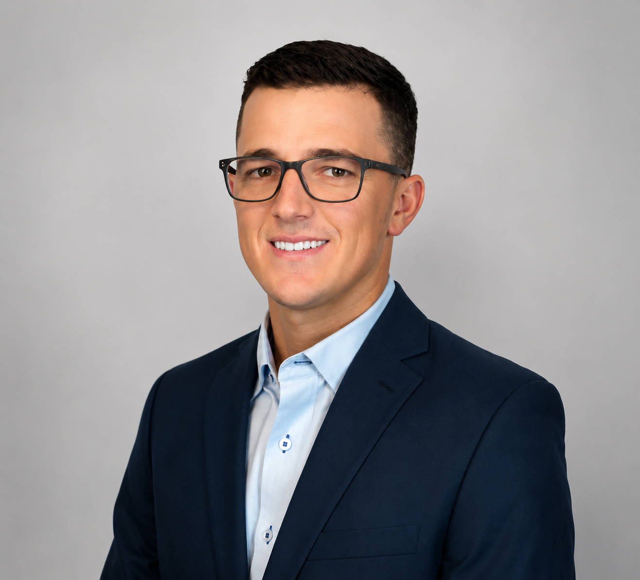 Professional headshot of a young man wearing glasses, a navy blazer, and a light blue shirt against a plain gray background.