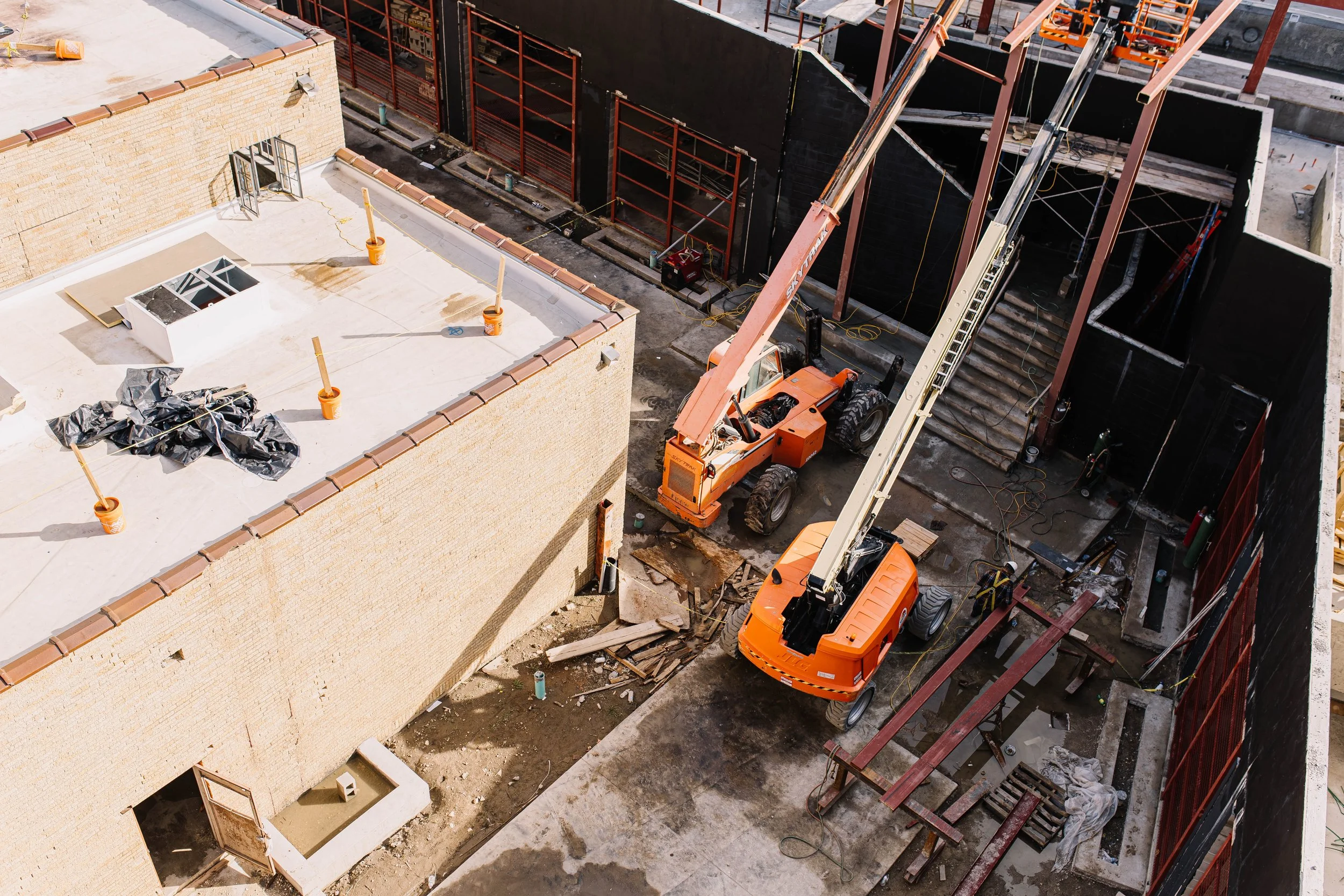 Construction site with cranes and partially built structures. A building with a beige brick exterior and open windows is visible in the foreground. The background shows more construction materials and equipment.