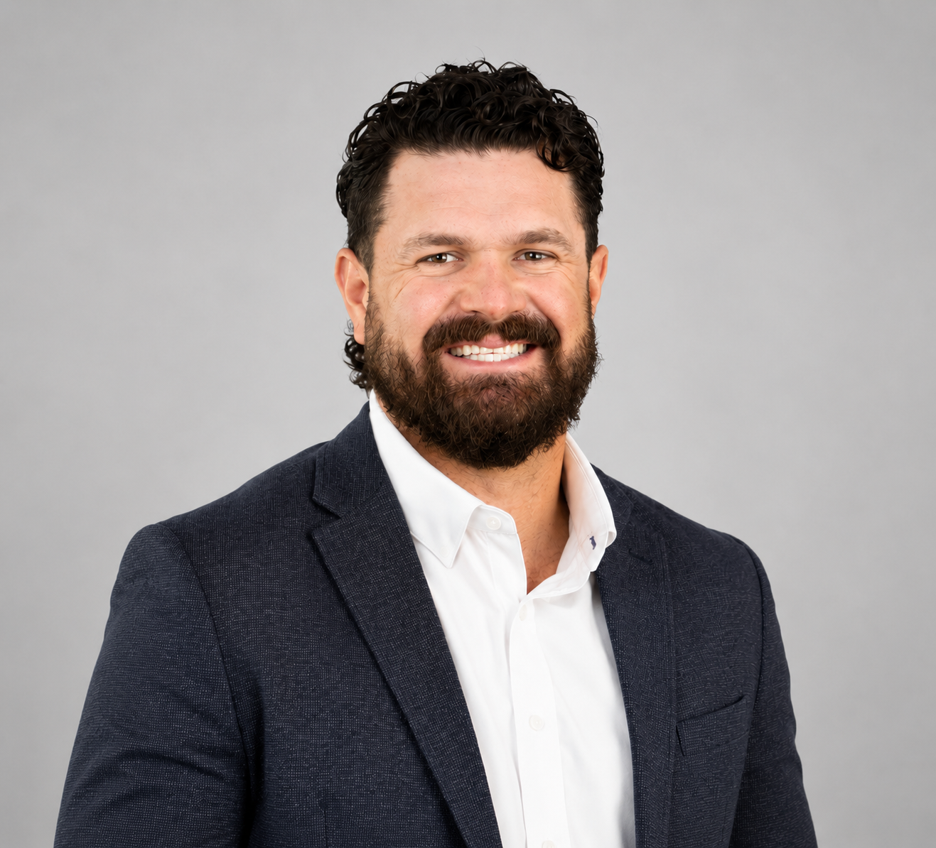 A man with dark curly hair and a full beard smiling, dressed in a white shirt and dark blazer, against a neutral gray background.