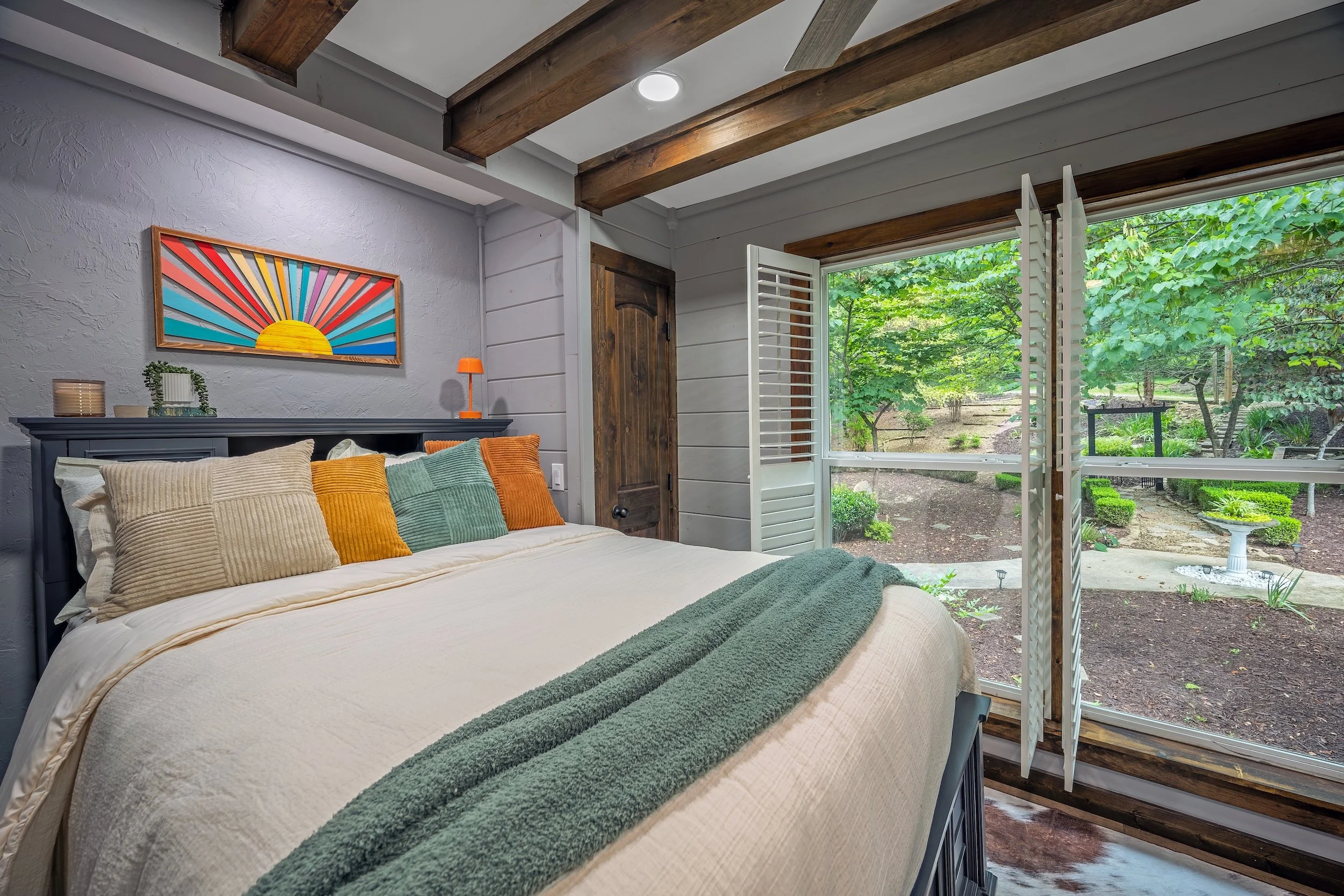 A cozy bedroom with a bed adorned with beige and green pillows, a dark gray headboard, and a large window overlooking a lush green garden with trees and shrubs. The room has white clapboard walls, wooden ceiling beams, and colorful wall art featuring a sunburst design.