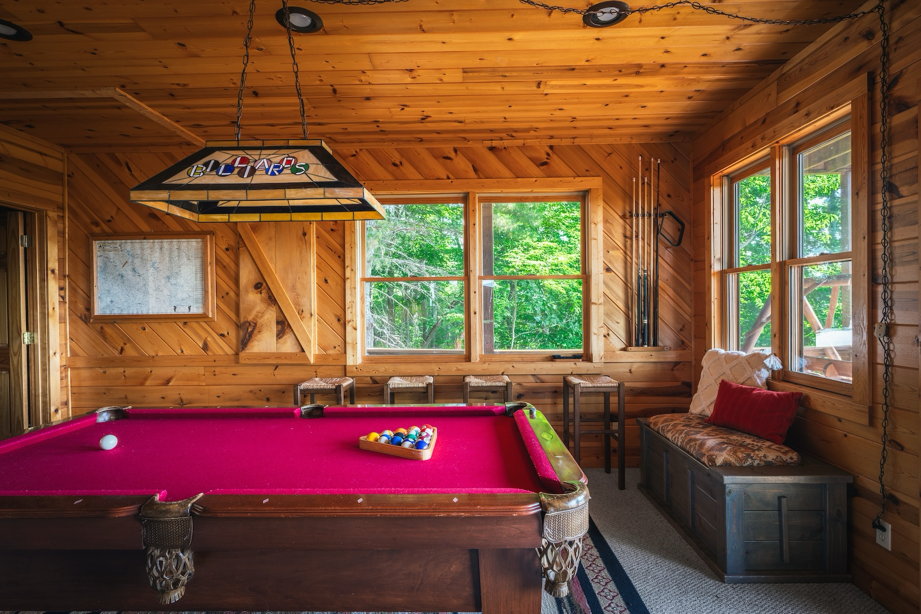 Interior of a wood-paneled game room with a pink pool table, cue sticks, a bench with cushions, and windows showing green trees outside.