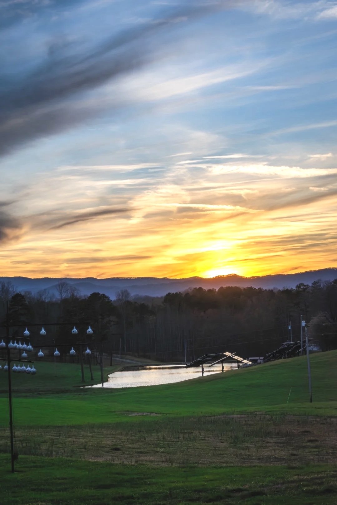 Sunset over a scenic landscape with mountains, trees, a pond, and a grassy field. Power lines and spherical lamps are visible.