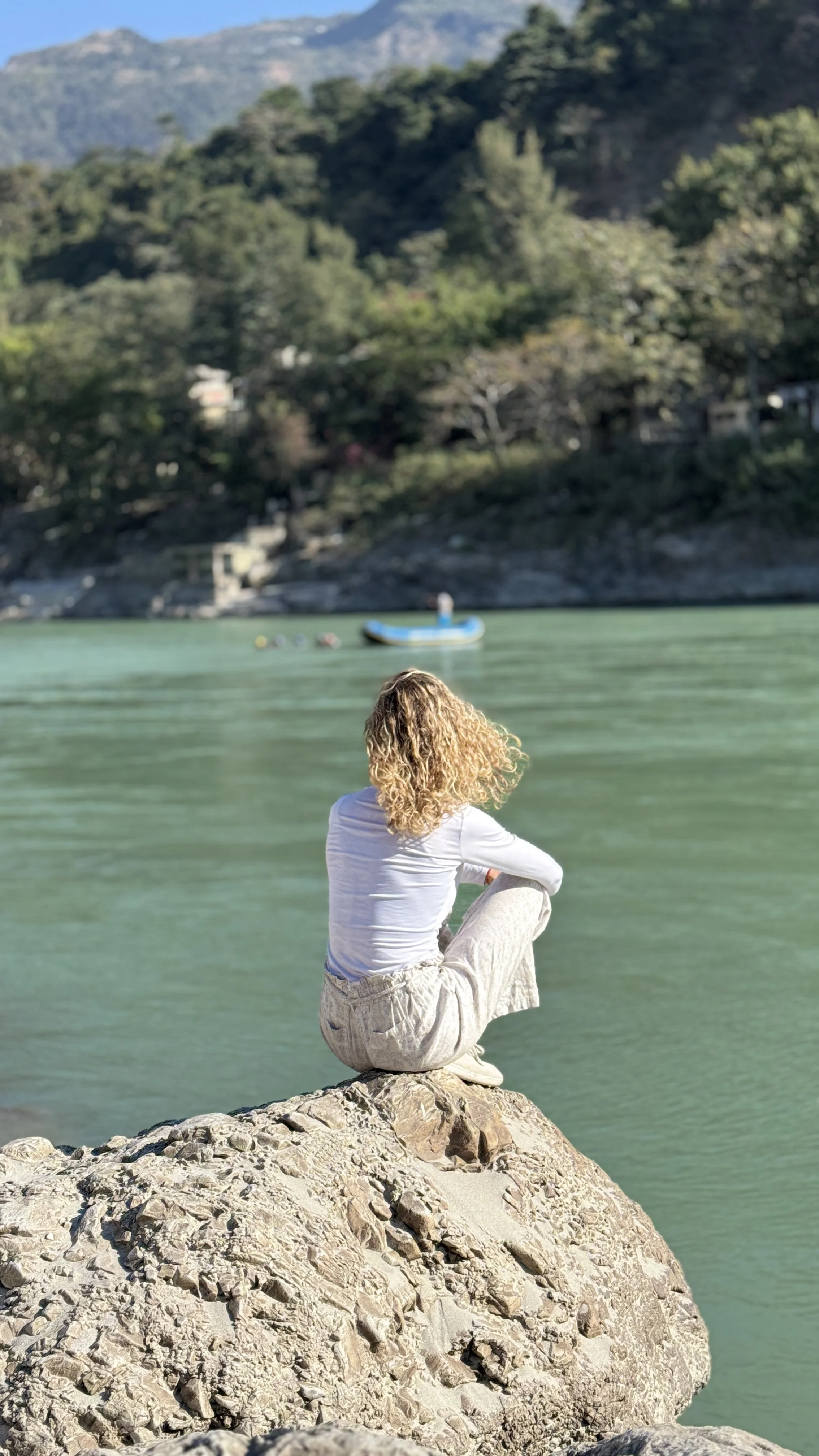 A person with curly blond hair sitting on a large rock by the water, facing away, looking at the river and mountainous landscape in the background.
