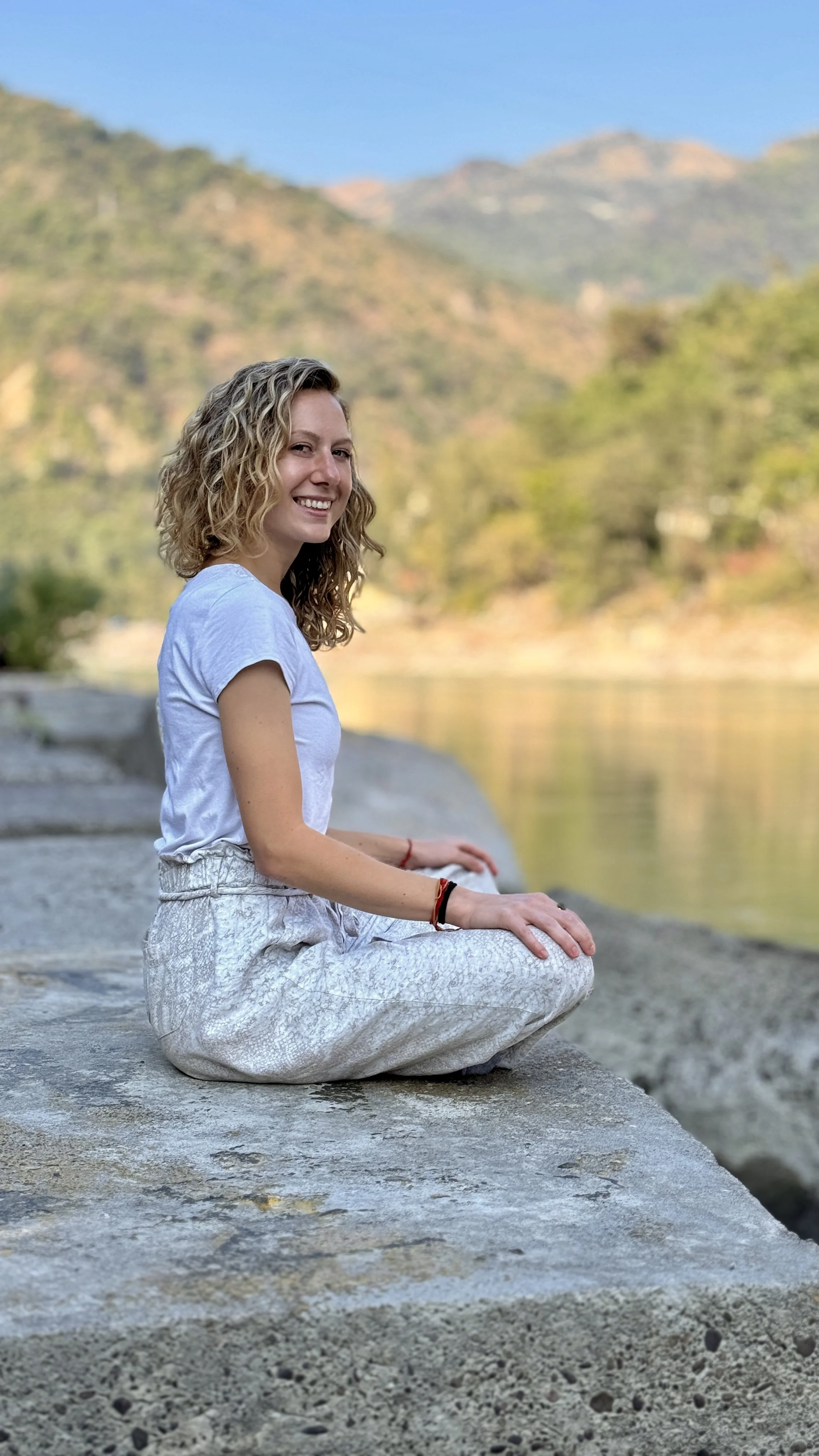 A young woman with curly hair, wearing a white T-shirt and patterned pants, sitting cross-legged on rock by a river with mountains and trees in the background.