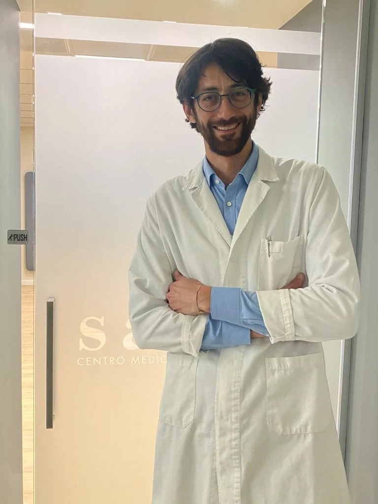 A man in a white lab coat and blue shirt standing with arms crossed, smiling, standing in front of a frosted glass door at a medical center.