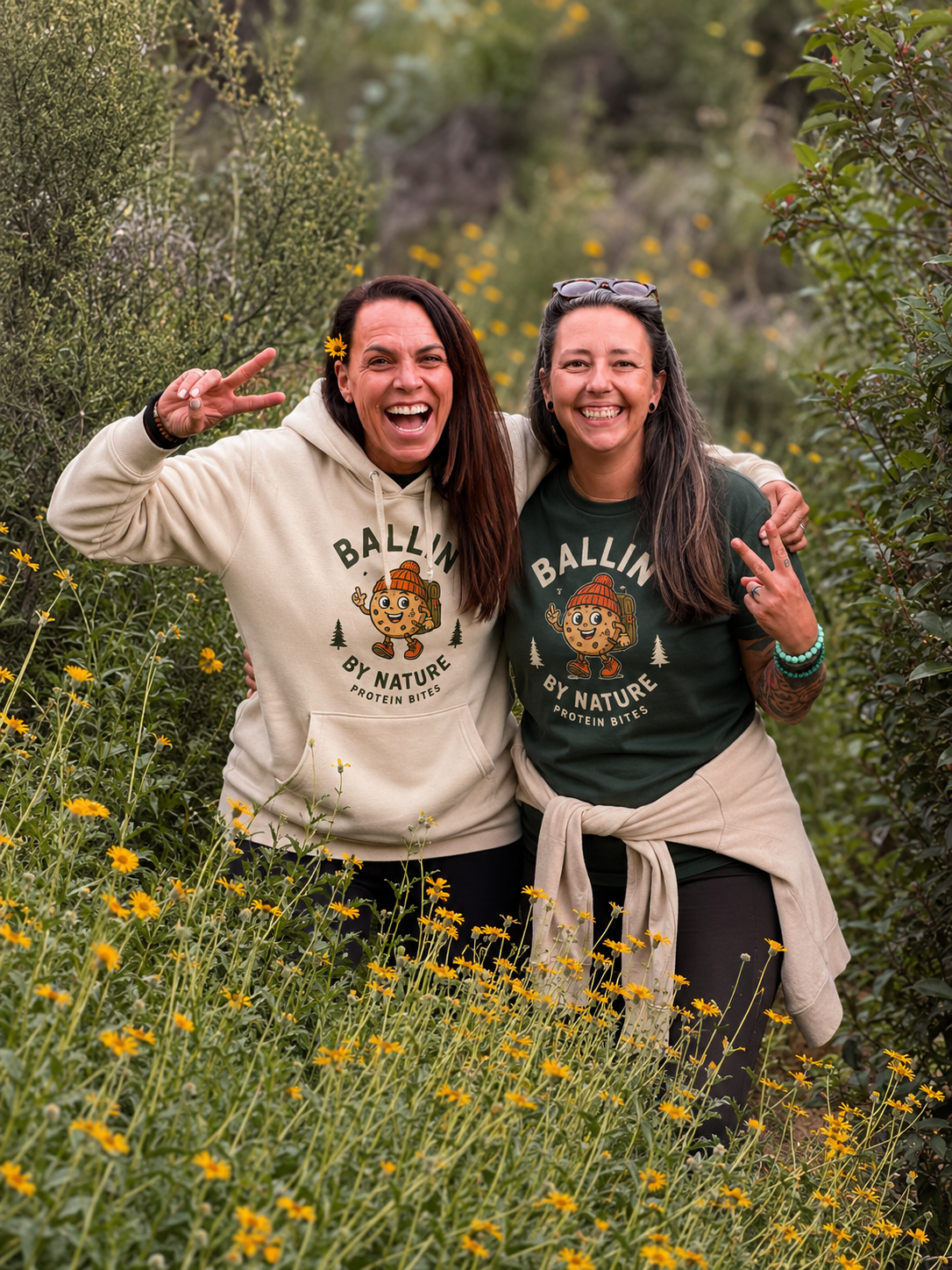 Two women smiling and making peace signs during a hike in a rocky, yellow wildflower-covered natural setting, wearing matching T-shirts with a cartoon character and the text 'Ballin' by Nature Protein Bites'.