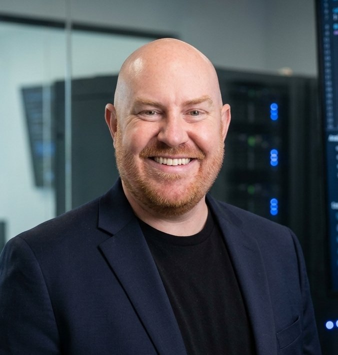 Portrait of a smiling man with a bald head and beard in a professional setting, wearing a black shirt and blazer, with server racks in the background.