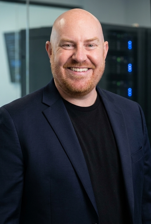 Man with a bald head and a red beard, smiling, wearing a dark blazer and black shirt in a server room with visible server racks.