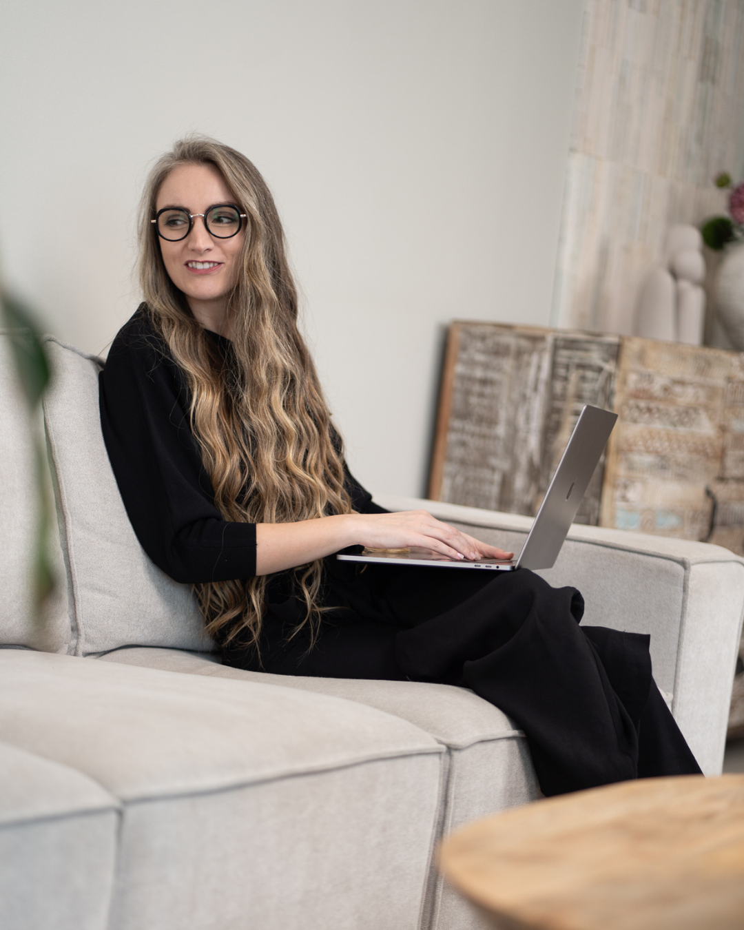 Woman with long wavy hair and glasses sitting on a beige couch, using a laptop.