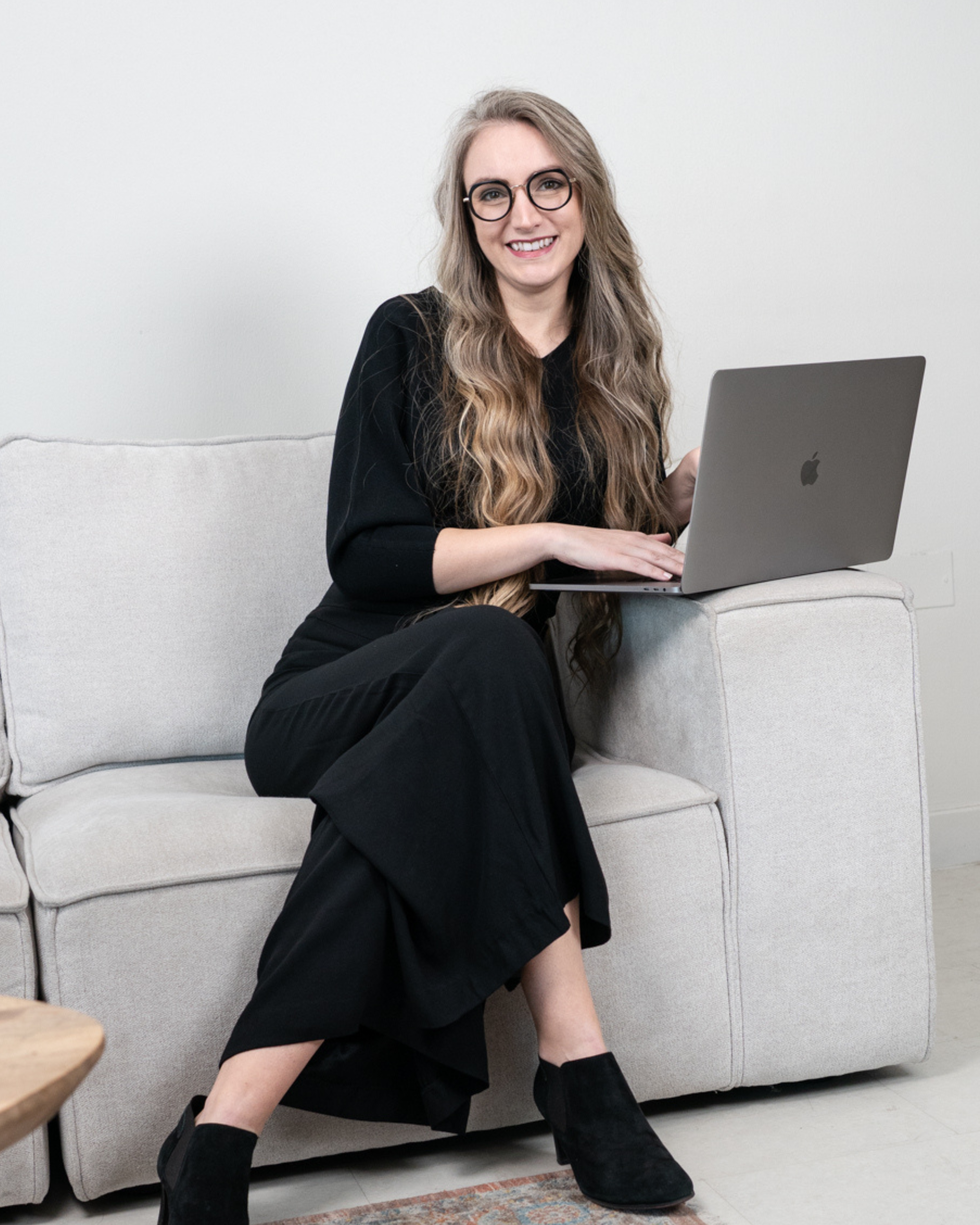 Martina Kikic wearing black shirt and pants sitting on sofa with laptop opened and smiling at camera. She has long curly hair. Promoting LinkedIn Live Workshop Masterclass.