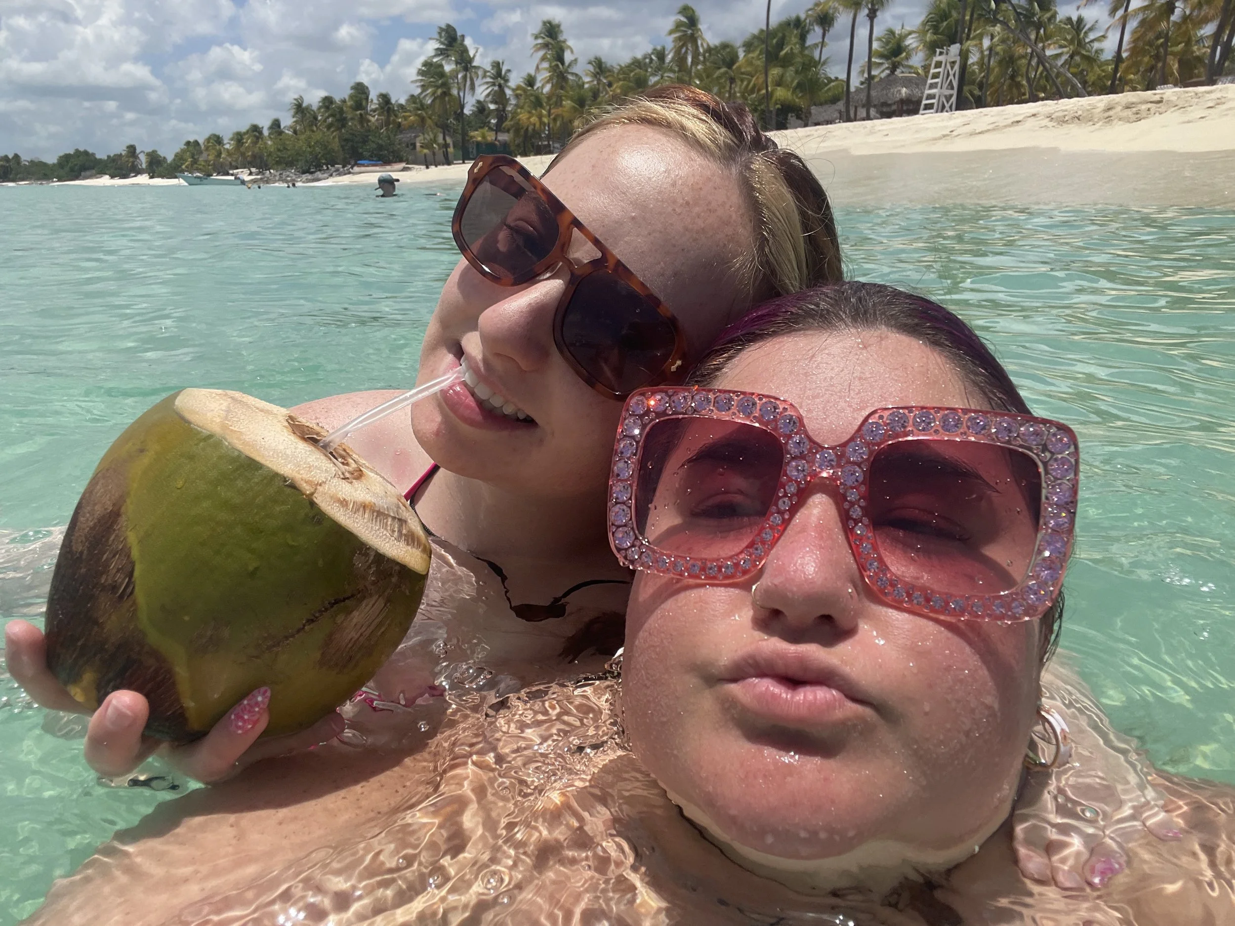 Two women at the beach, one with sunglasses drinking from a coconut with a straw, and the other with sunglasses covered in rhinestones taking a selfie in clear water with palm trees and a sandy beach in the background.