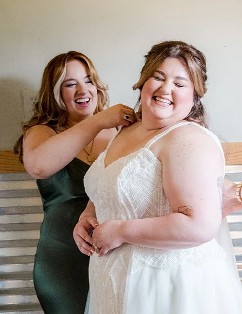 Two women smiling, one in a white wedding dress, being helped with her dress by another woman in a dark dress.