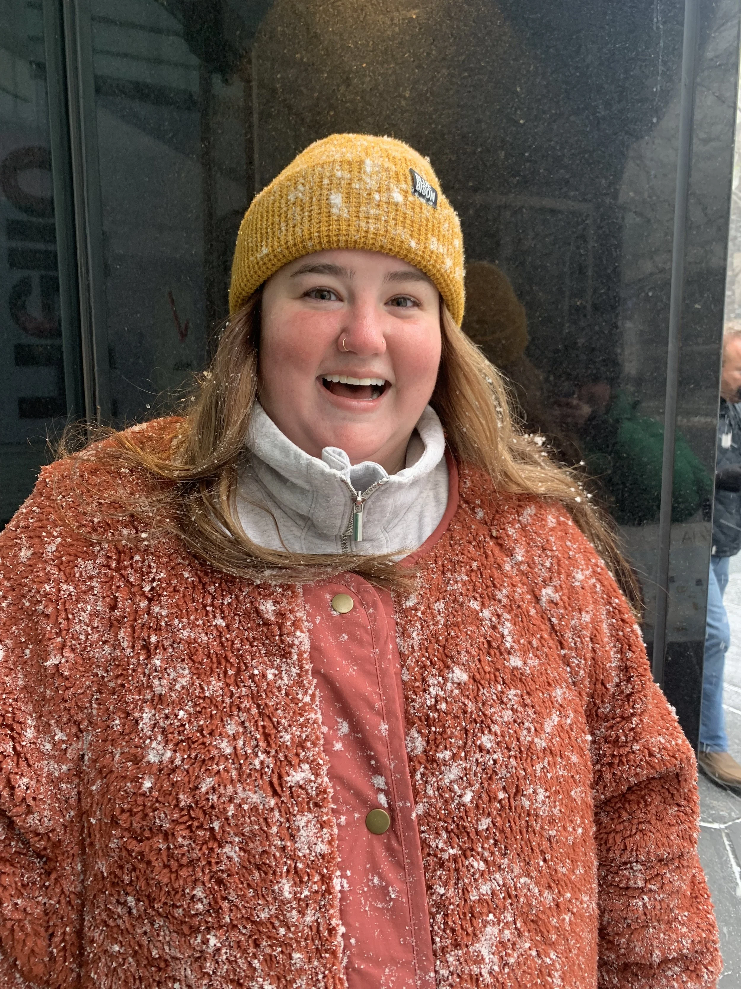 A smiling young woman with long reddish-brown hair wearing a yellow knit beanie with snow on it, a gray zip-up jacket, and a coral-colored textured coat covered in snow, standing near a glass elevator or window with a dark background.