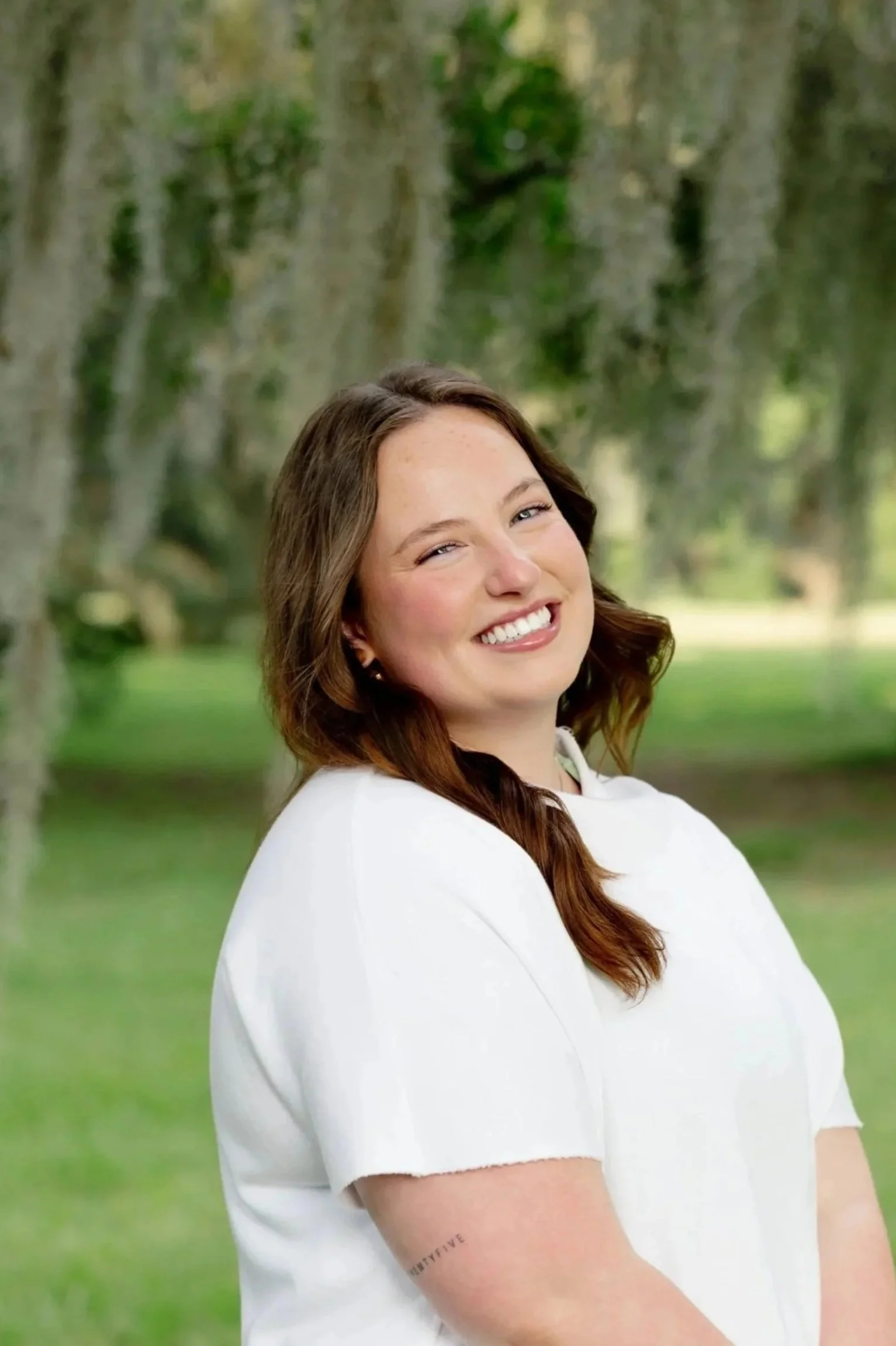 A woman with long brown hair smiling outdoors in front of greenery, wearing a white top and showing a bracelet on her arm.