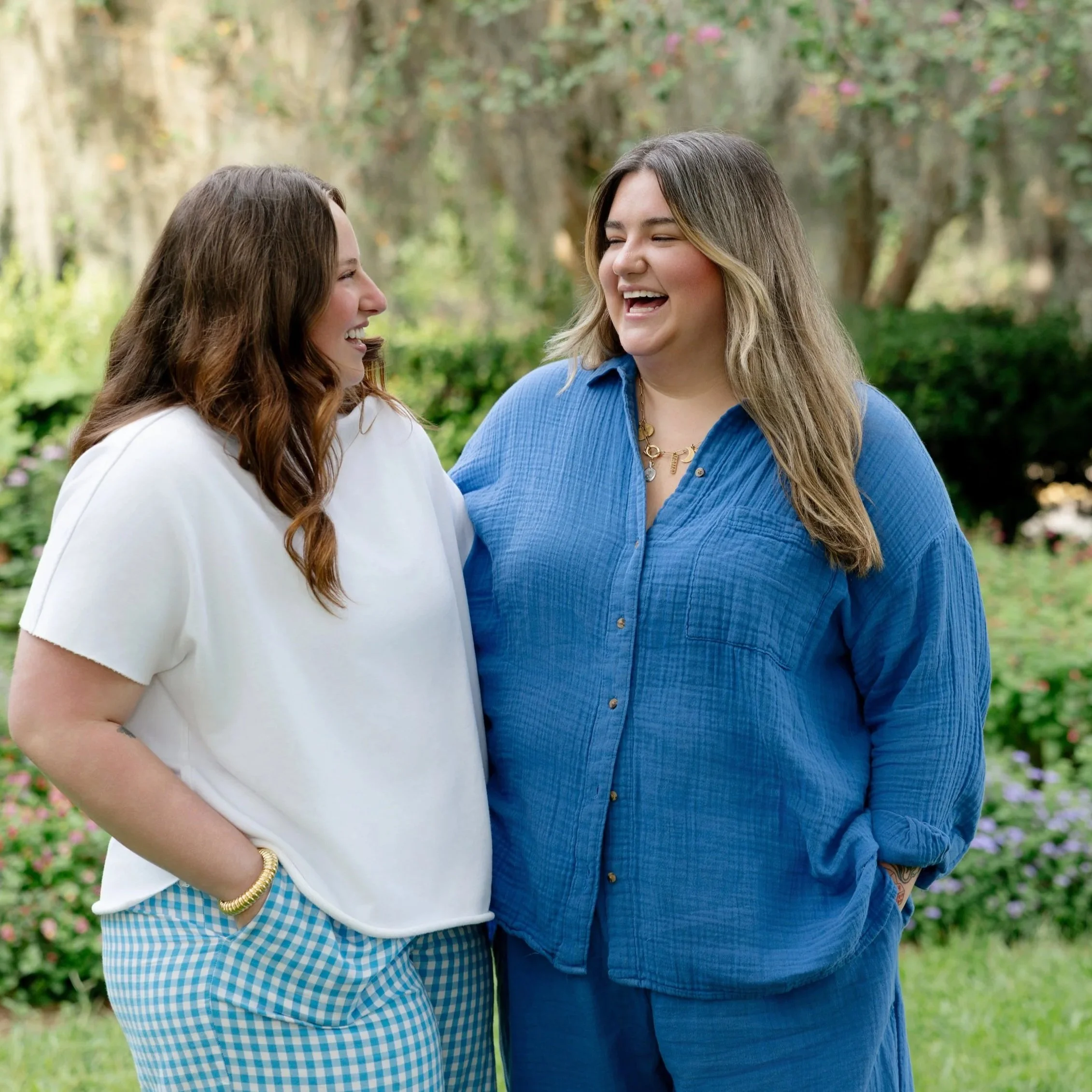 Two women standing outdoors, smiling and laughing with each other, in a park or garden setting with trees and greenery in the background.