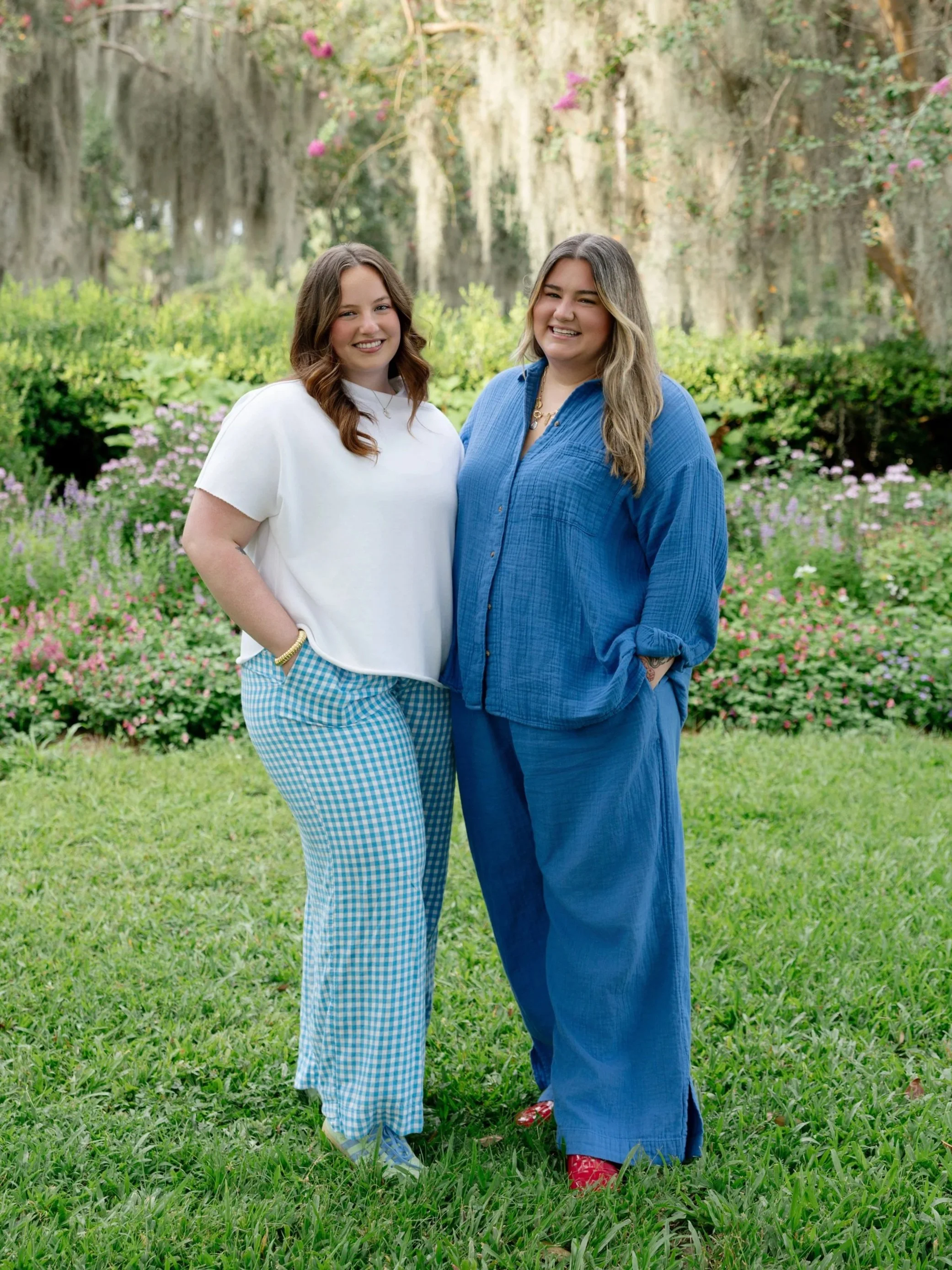 Two young women standing together outdoors, smiling, with greenery and hanging moss in the background.
