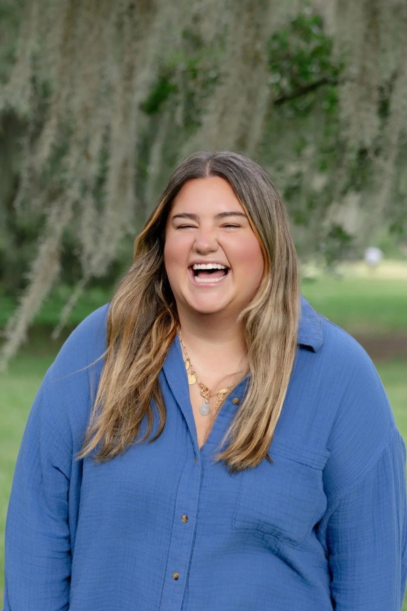 A woman with long, light brown hair smiling outdoors in a park, wearing a blue button-up shirt and gold jewelry.