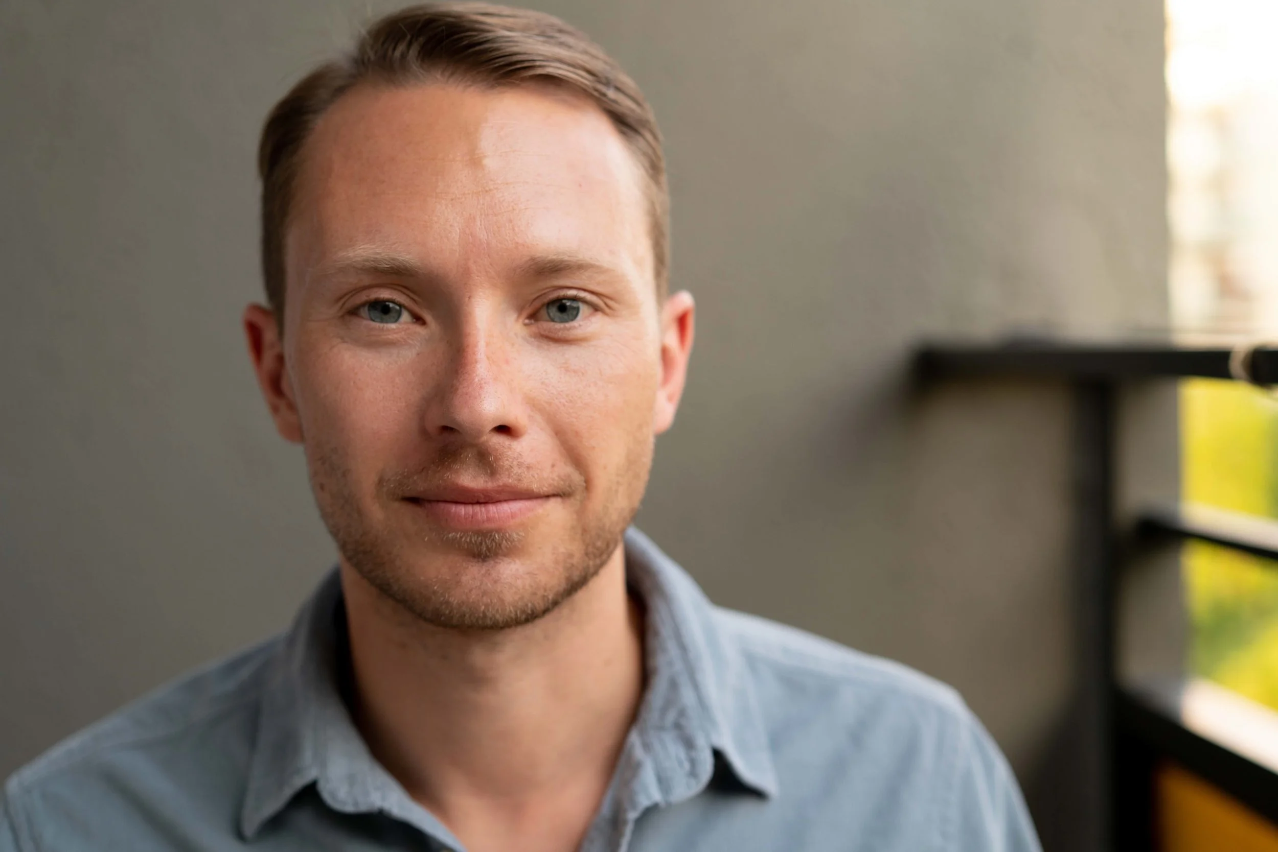 Close-up of a young man with light brown hair and blue eyes, smiling slightly, wearing a light blue collared shirt, standing against a neutral background with a window and shelves.
