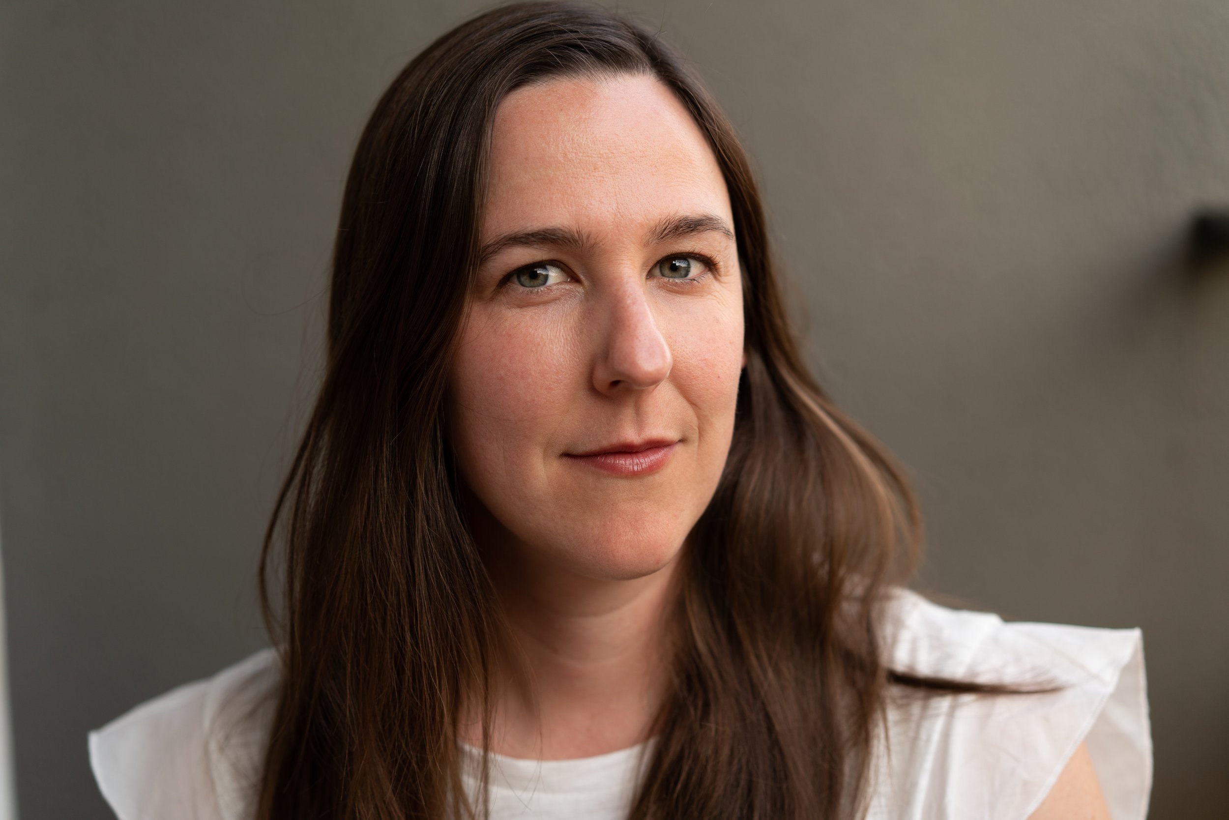 Close-up of a woman with long brown hair and gray eyes, wearing a white top, against a gray background.