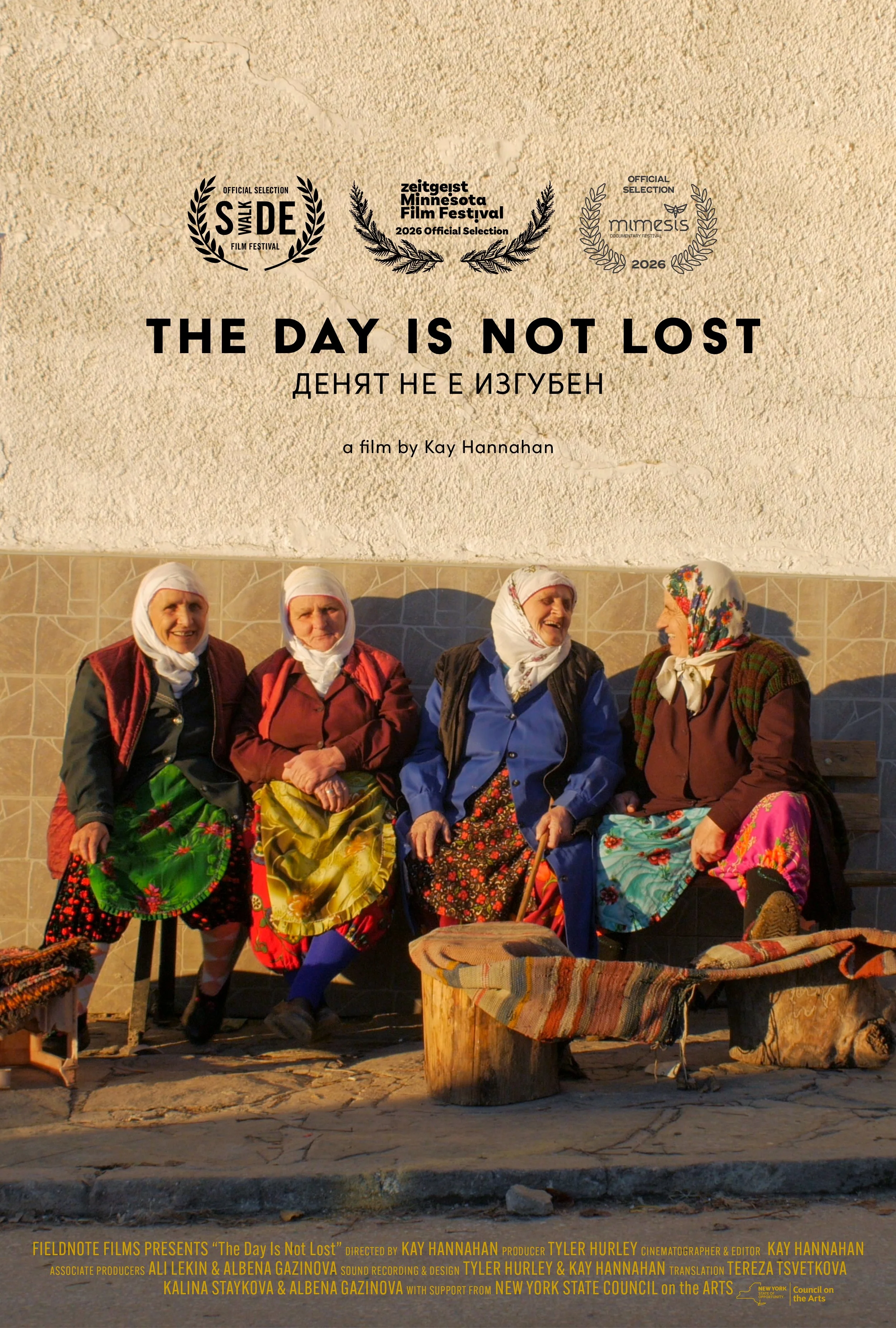 Four elderly women in traditional clothing sit and stand in front of a beige wall with a textured surface. They are smiling and engaging with each other, sitting on chairs and a wooden bench with a wooden table in front of them, which is covered with a colorful woven cloth. The image is part of a promotional poster for a film titled 'The Day Is Not Lost' by Kay Hannahan.