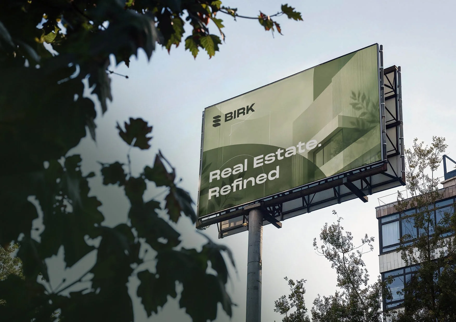 A large outdoor billboard with a green geometric design displaying the words 'BIRK' and 'Real Estate. Refined.'. The billboard is supported by a tall pole, with trees and a modern building with glass windows visible in the background, and the sky appears overcast.