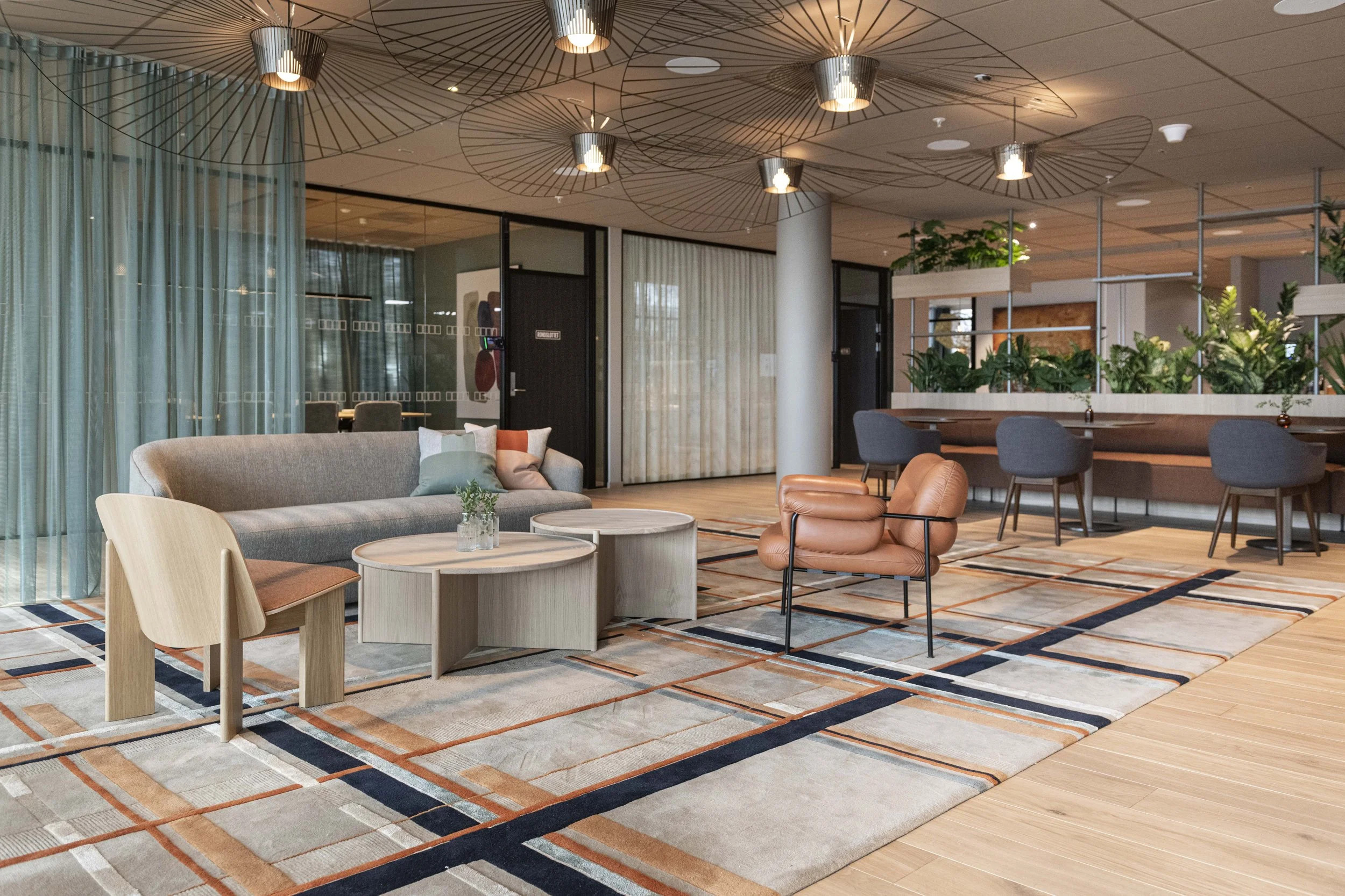 Modern hotel lobby with seating area, decorative ceiling lights, a patterned area rug, and indoor plants.