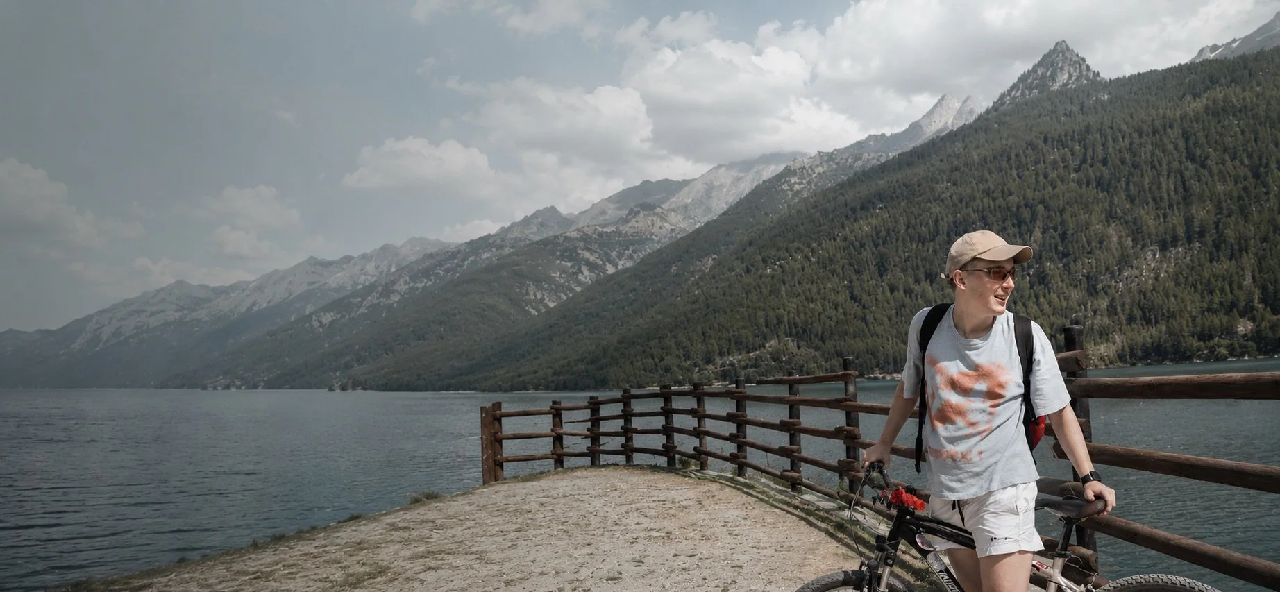 A person with short hair, wearing sunglasses, a tan cap, a white t-shirt with orange and gray design, white shorts, holds a bicycle, and stands on a gravel path by a wooden railing near a lake surrounded by mountains and forests on a cloudy day.
