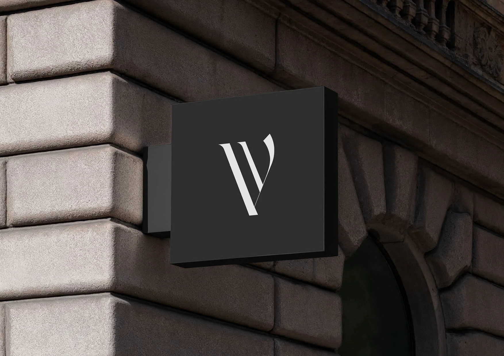 Close-up of a black sign with a stylized white letter "V" mounted on a beige brick wall.