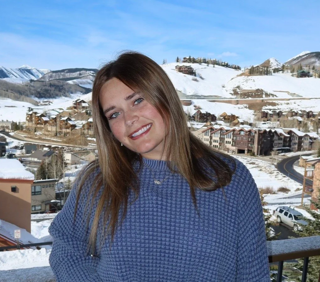 A woman with light brown hair and blue eyes smiling outdoors on a snowy mountain village with houses and ski slopes in the background.