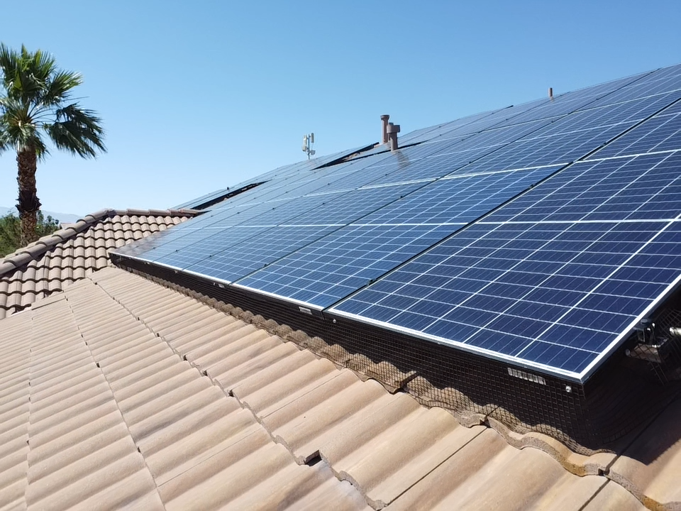 Residential roof with multiple solar panels installed on top, a palm tree and a blue sky in the background.