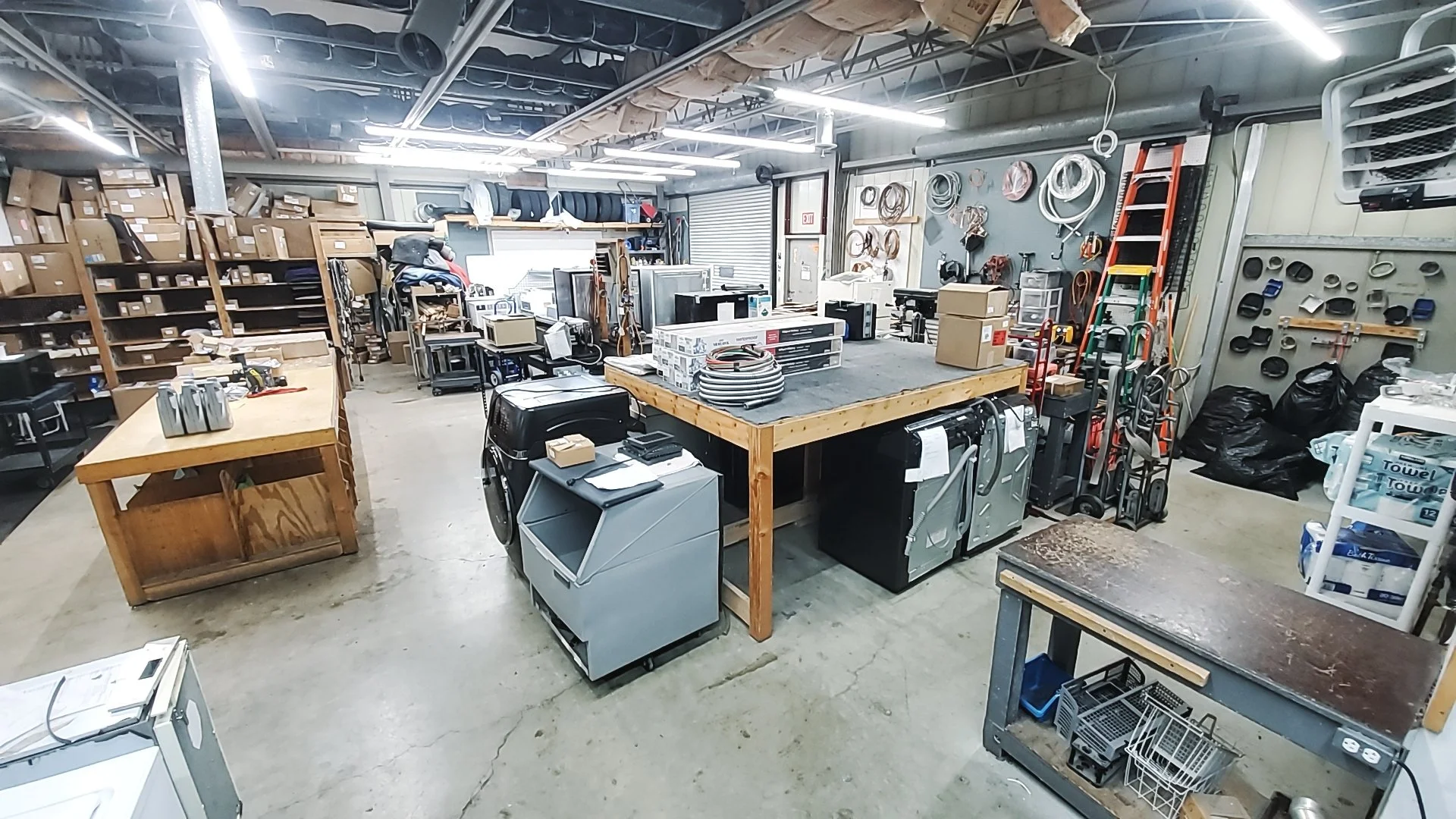 A workshop or storage room filled with various tools, equipment, and supplies. There are shelves with boxes, cutting tools, hoses, ladders, and bins.