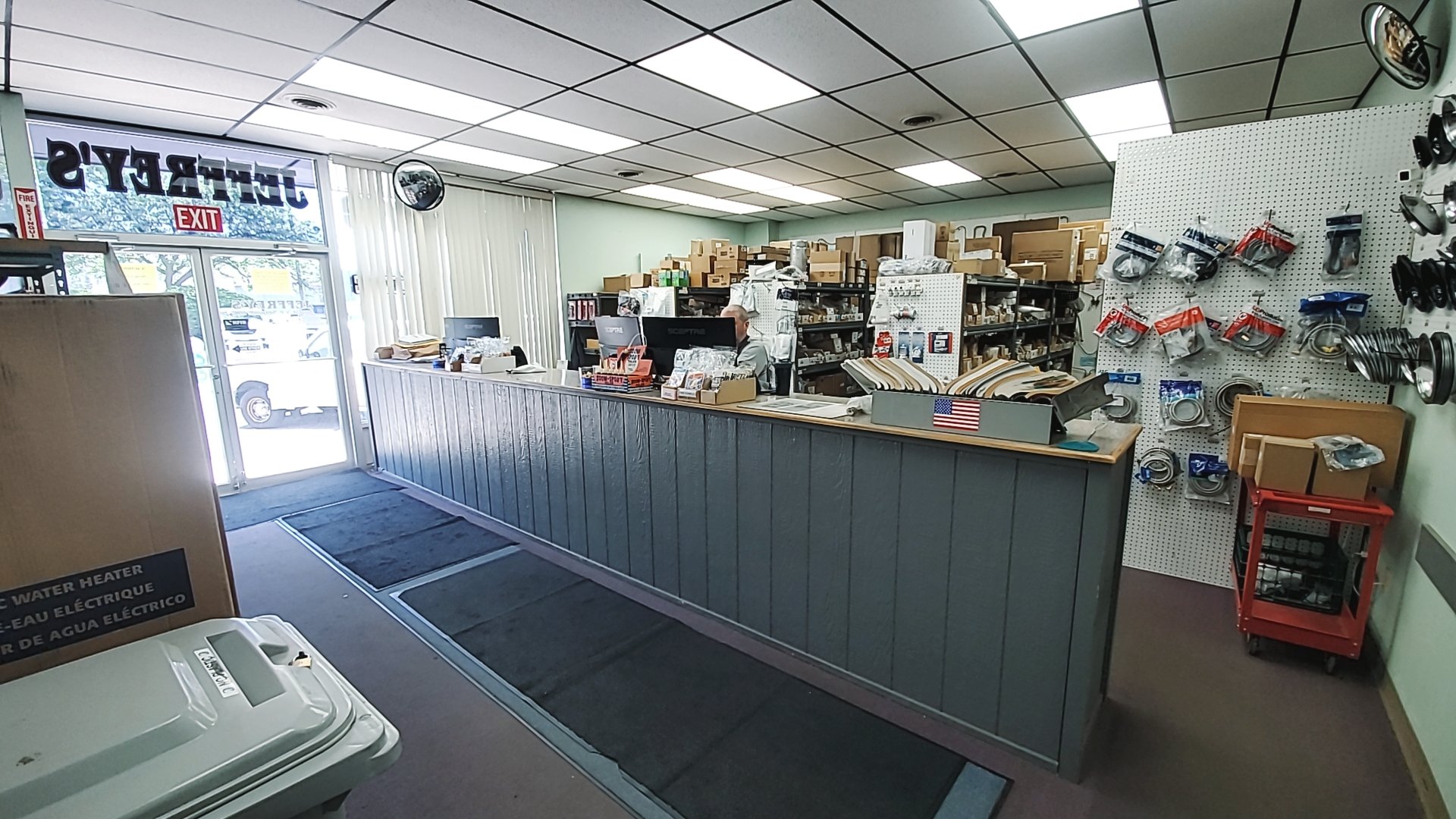 Interior of a shipping or hardware store with a counter, shelves filled with boxes, tools, and accessories, a customer service representative working at a desk, and an exit door with a glass panel showing outside parking lot and trees.