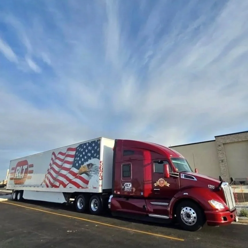 A large red semi-truck with an American eagle and flag design on the trailer, parked in a lot next to a beige building.