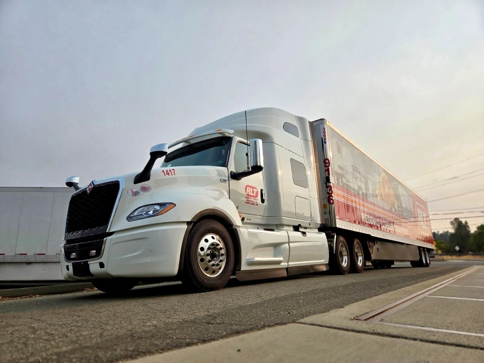 A white semi-truck with a red trailer parked on the side of a road during sunset.